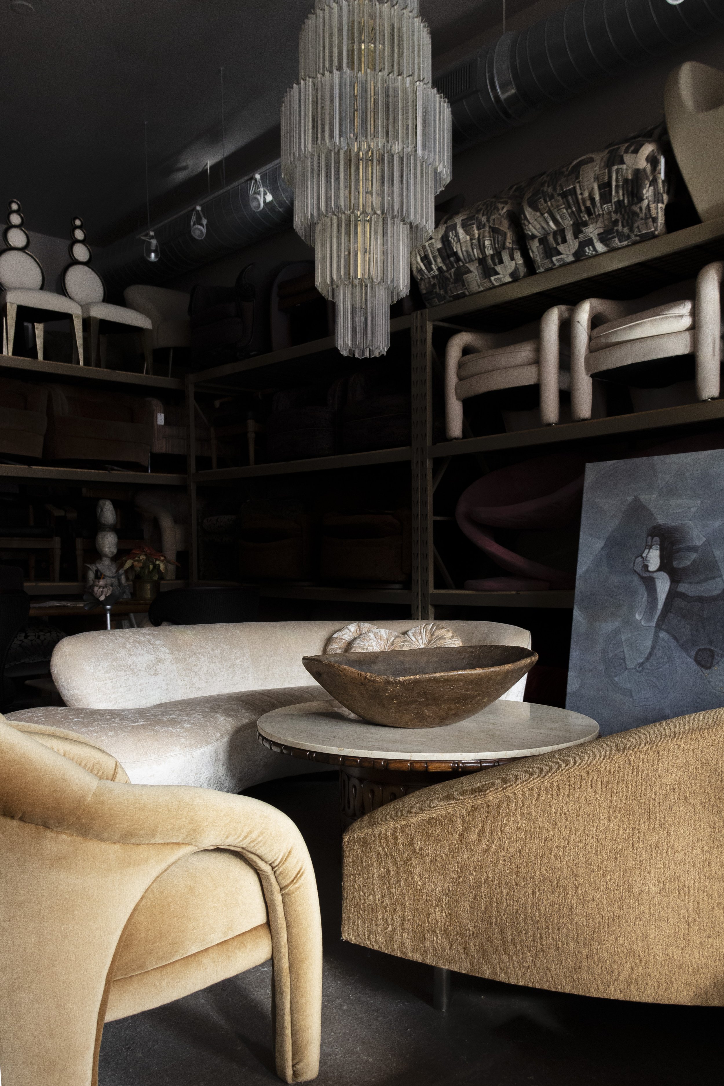 A vintage-style interior with a crystal chandelier, a round wooden table, beige curved sofas, and an abstract painting. Shelves in the background display stacked furniture pieces.