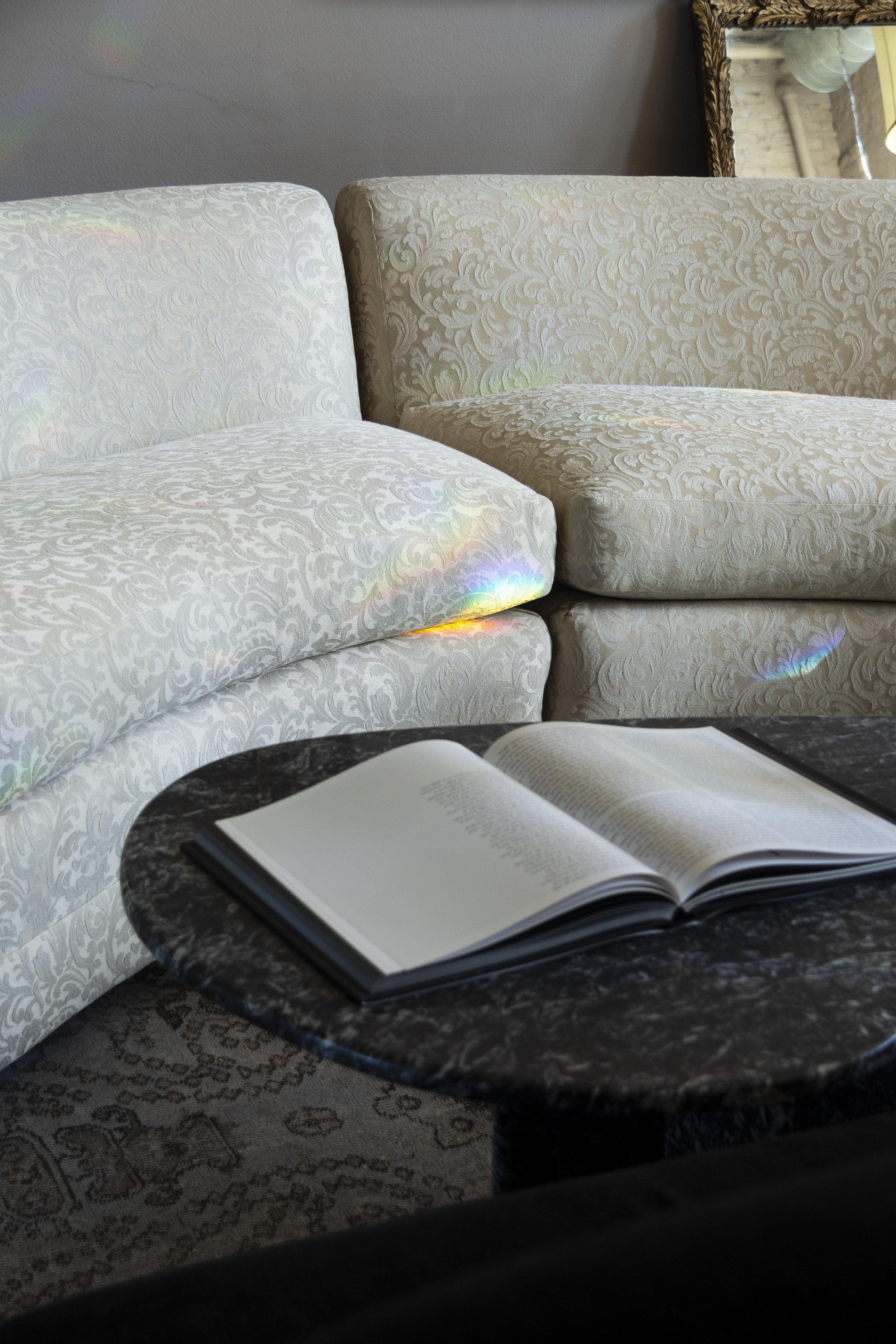 Open book on a black marble coffee table with patterned beige couches in the background, illuminated by rainbow light reflections.