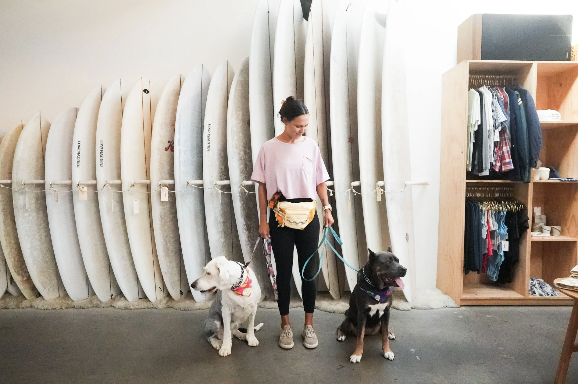 A woman with dark hair tied back, wearing a light pink t-shirt, black pants, and sneakers, holding leashes of two dogs in a surf shop. One dog is white with a black collar, and the other is black with brown markings and a colorful bandana. Behind them are racks of surfboards, and to the right, shelves with clothes and accessories.