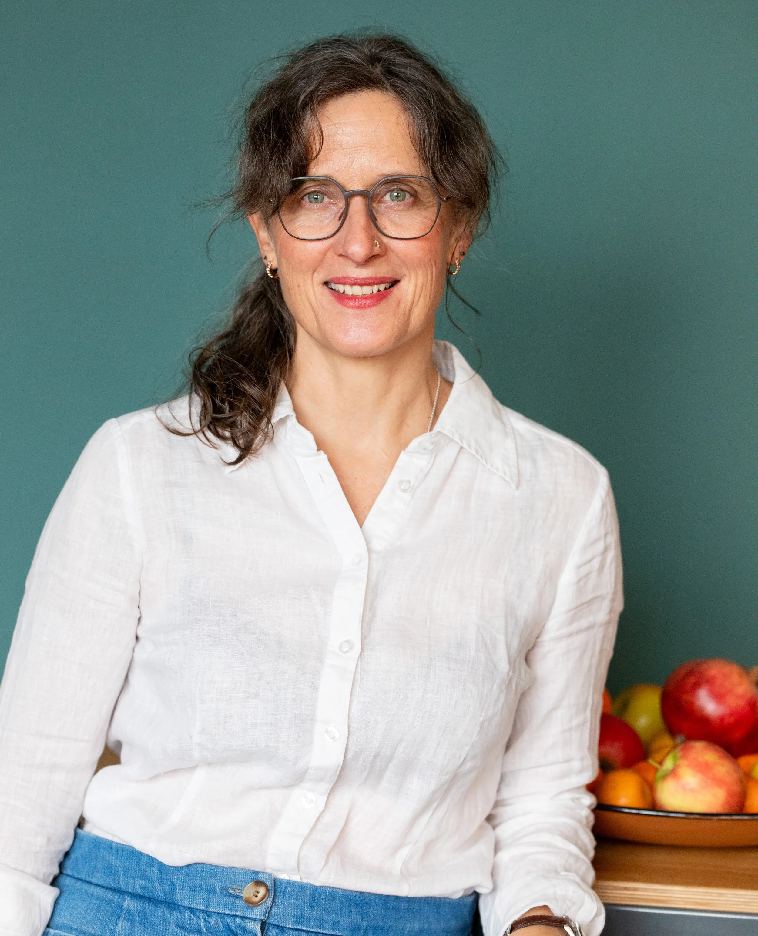 Julia Nowak, Ernährungsberaterin in Berlin, in weißem Hemd sitzt vor einem Teller mit Äpfeln vor einem grünen Hintergrund.