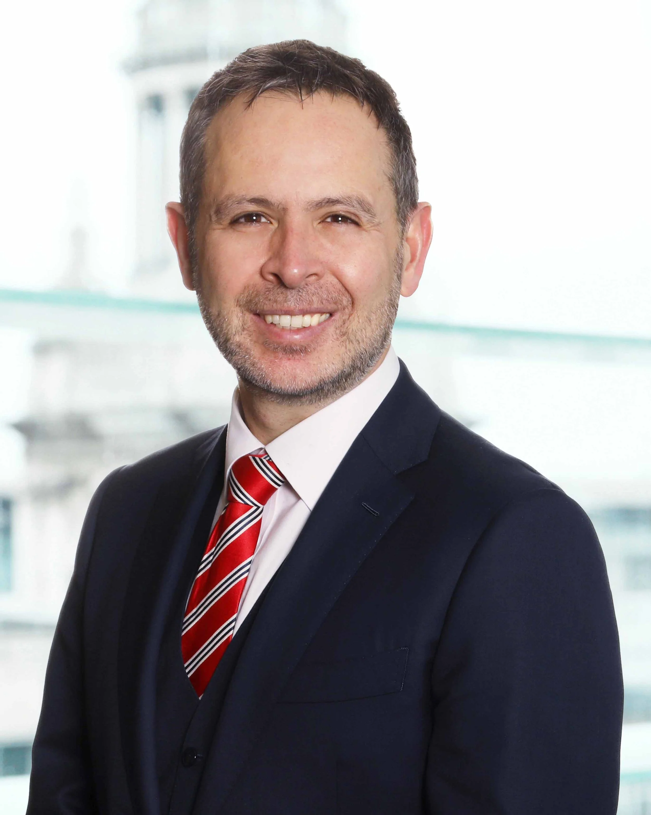 Portrait of a smiling man in a suit with a red, white, and blue striped tie, standing in front of a blurred office building background.