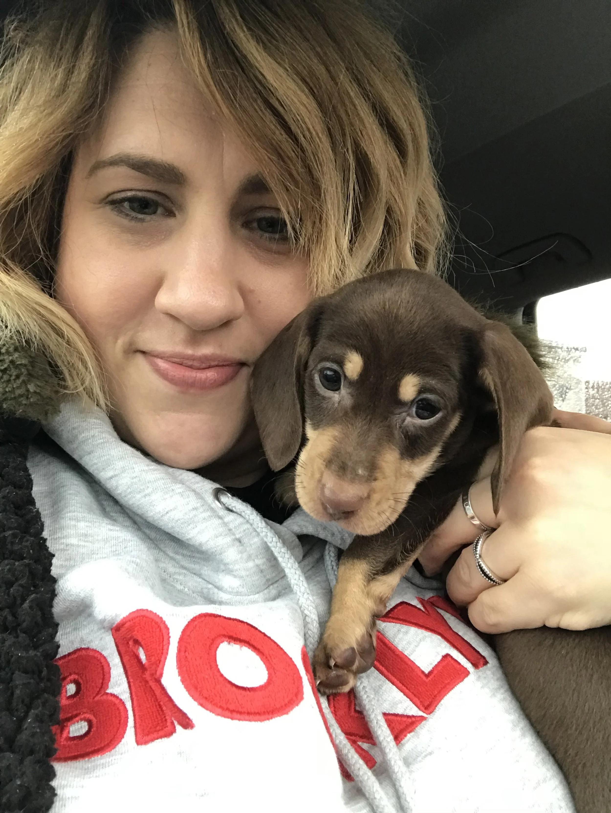 A woman with blonde hair holding a brown Dachshund puppy inside a car.