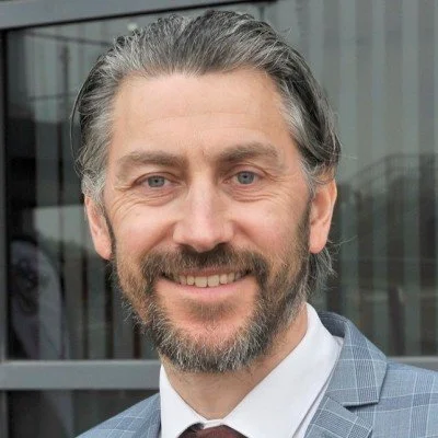 A smiling middle-aged man with gray hair and beard, wearing a suit and tie, standing outside a modern building.