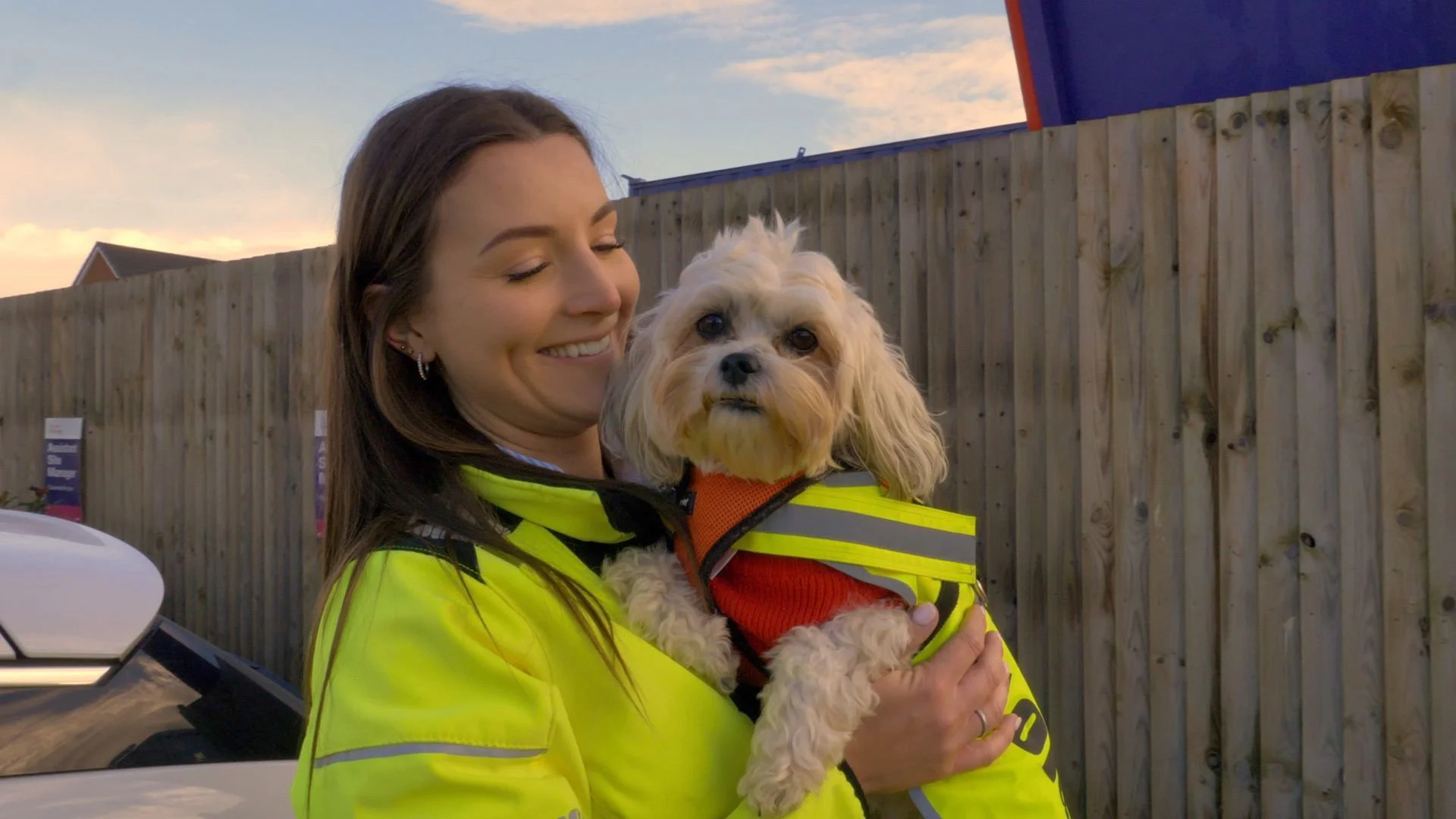 A woman in a high visibility yellow jacket smiling while holding a small beige dog in a similar yellow vest outside. The background shows a wooden fence and a partly cloudy sky at sunset.