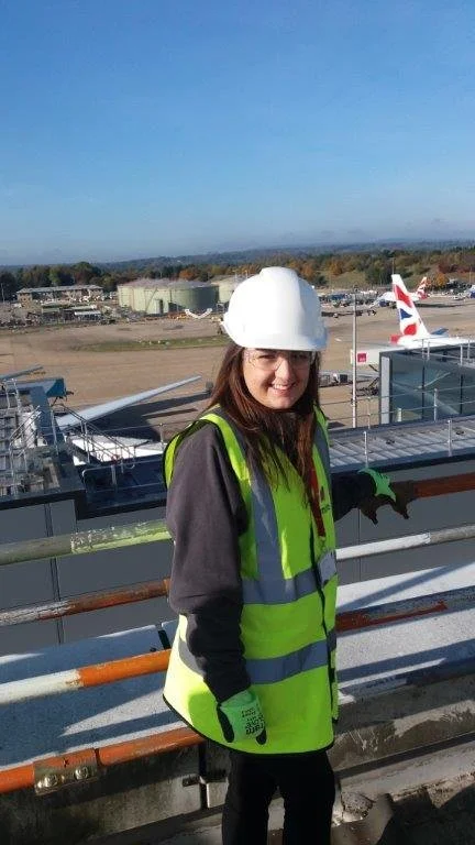 A woman wearing a white safety helmet and a high-visibility yellow safety vest standing on an airplane runway or observation deck at an airport, with commercial aircraft and airport infrastructure in the background.