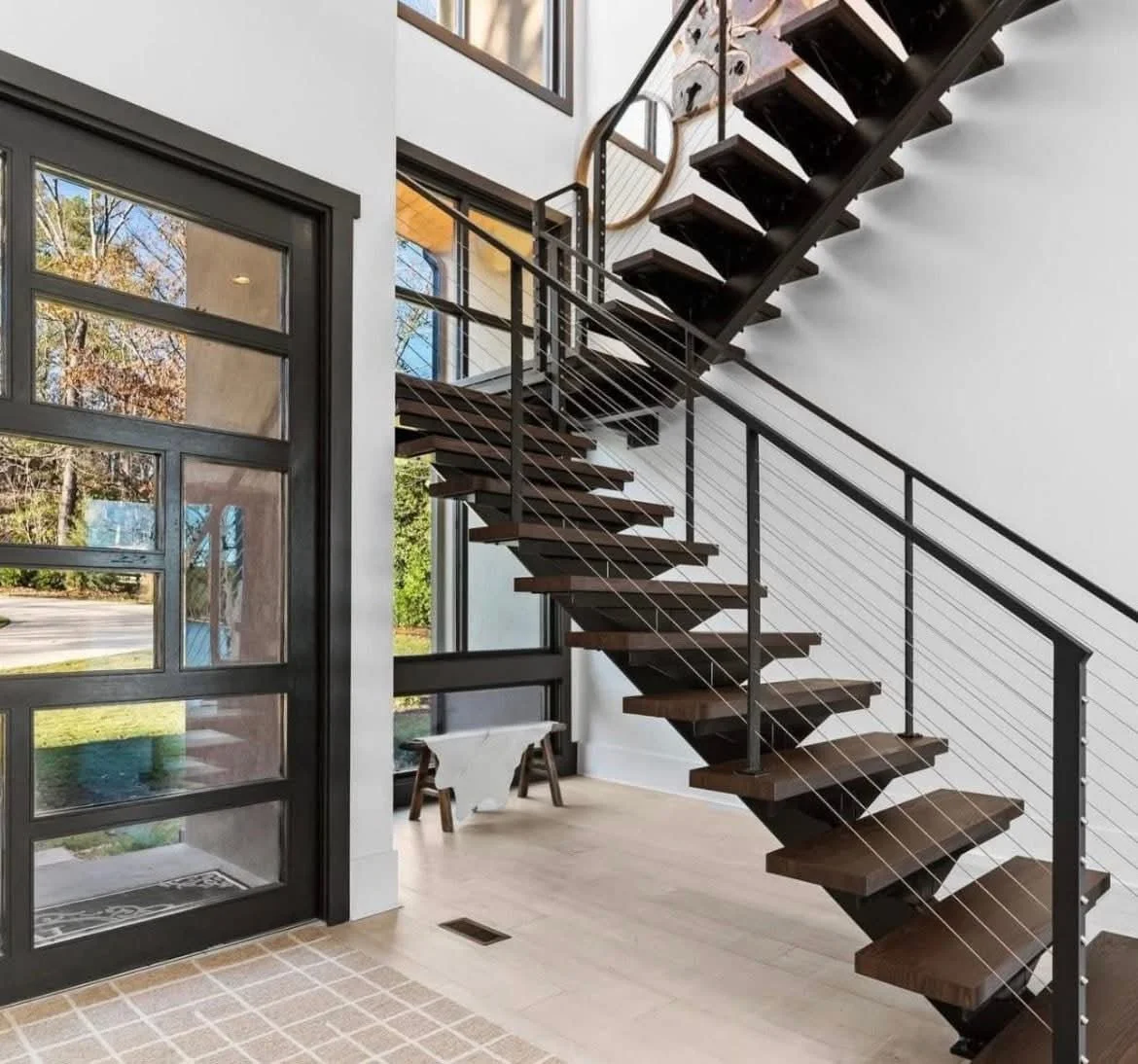 Interior view of a modern home with a floating staircase made of dark wood and metal, next to large glass windows and a black-framed door, with a doormat on a shiny tiled floor.