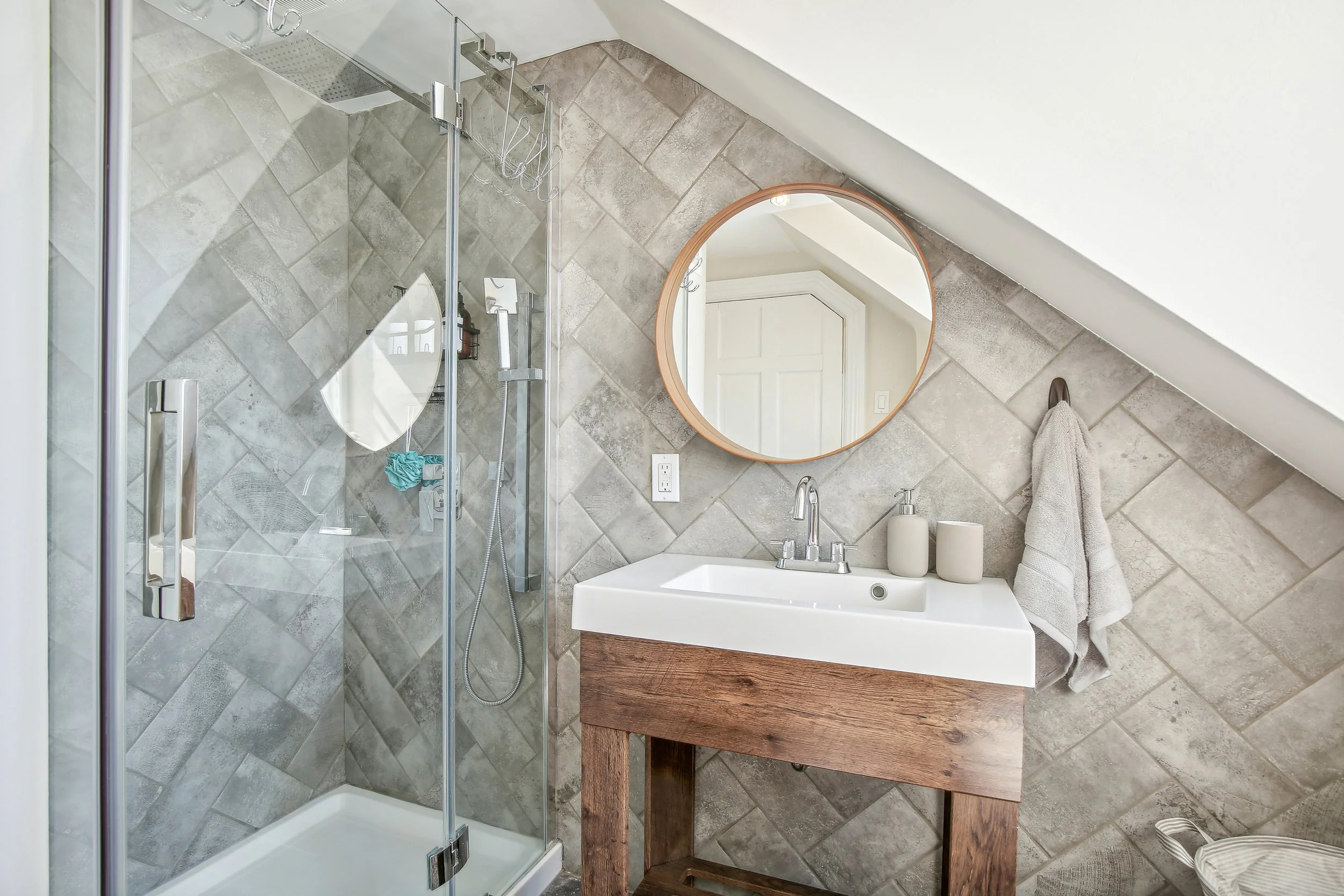 Bathroom with a glass shower enclosure, a round mirror, a white sink with wooden base, and beige tiled walls.