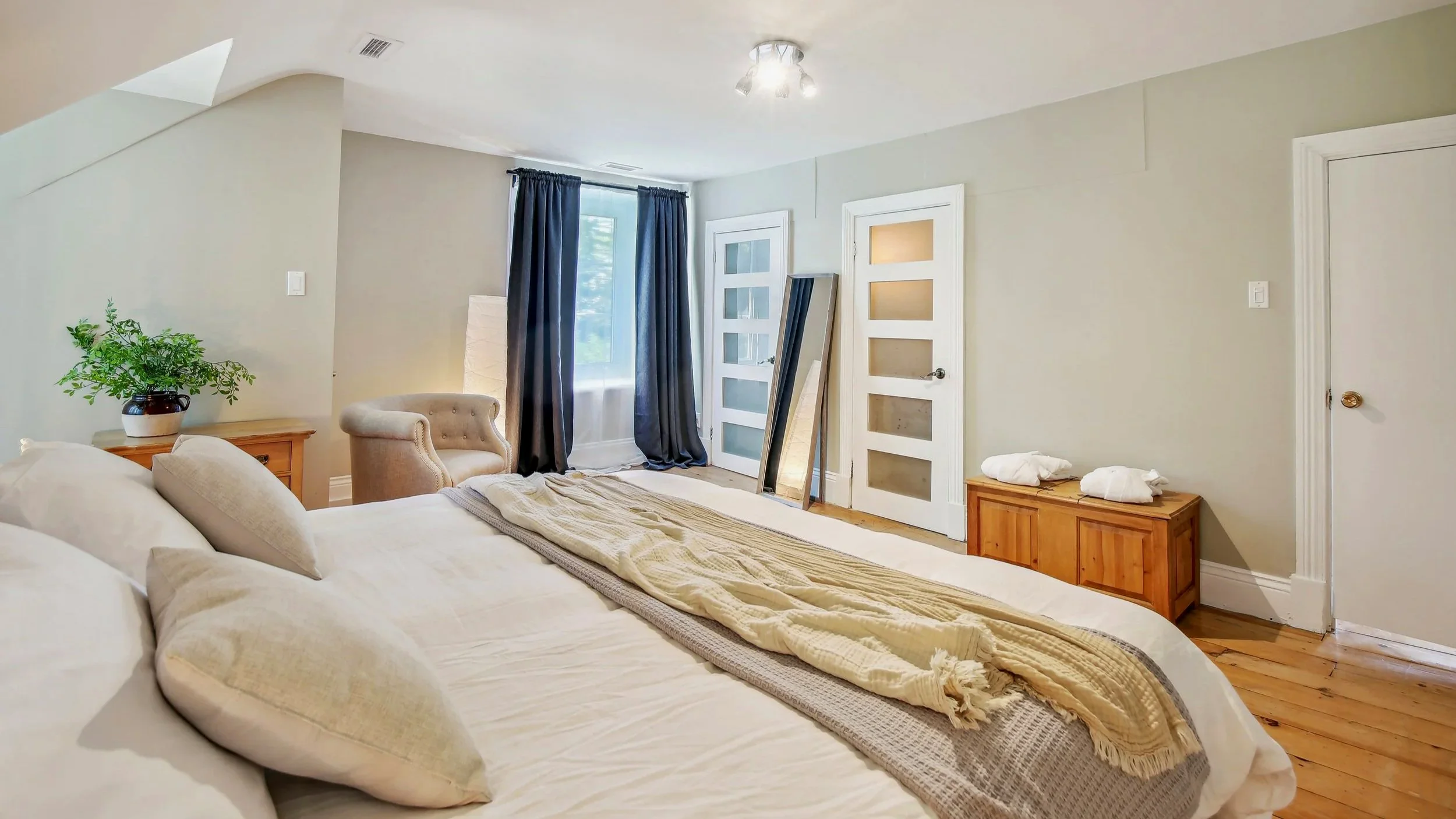 A bedroom with a bed, a beige armchair, a wooden side table with a plant, a dresser with two towels, and a window with dark curtains.
