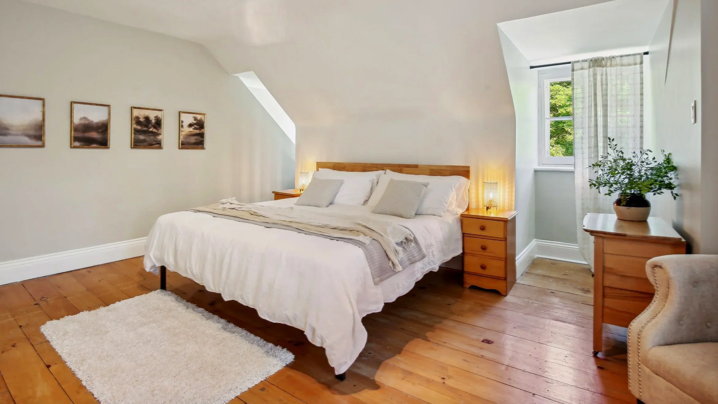 A cozy bedroom with a wooden bed, white bedding, and pillows. There are two nightstands with lamps, a window with sheer curtains, a potted plant, and a small dresser. The walls are decorated with four landscape paintings, and the floor is wooden with