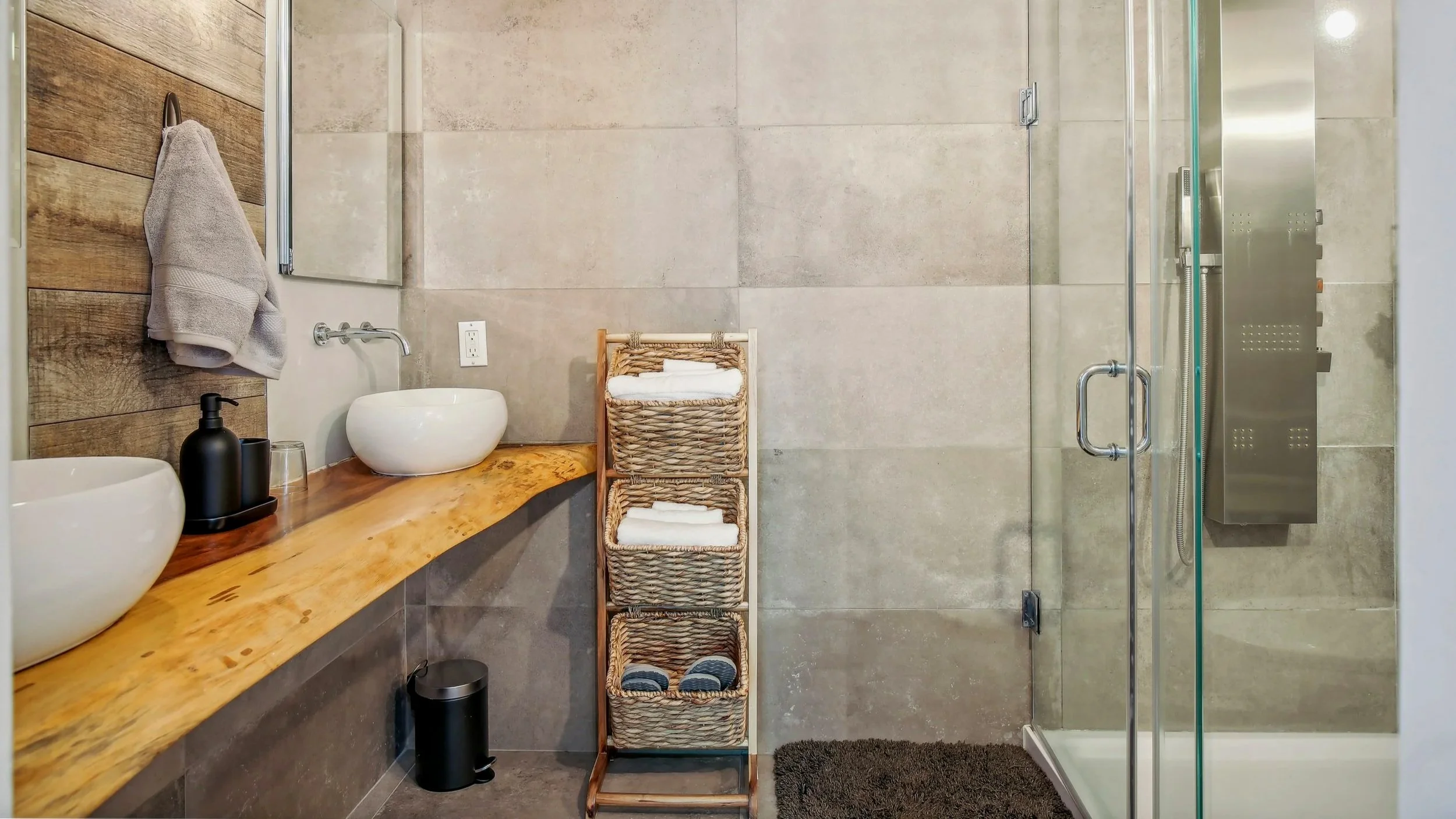Modern bathroom with dual vessel sinks on a wooden vanity, a wicker storage rack with towels, a glass shower enclosure with a stainless steel shower panel, and neutral-colored tiled walls.