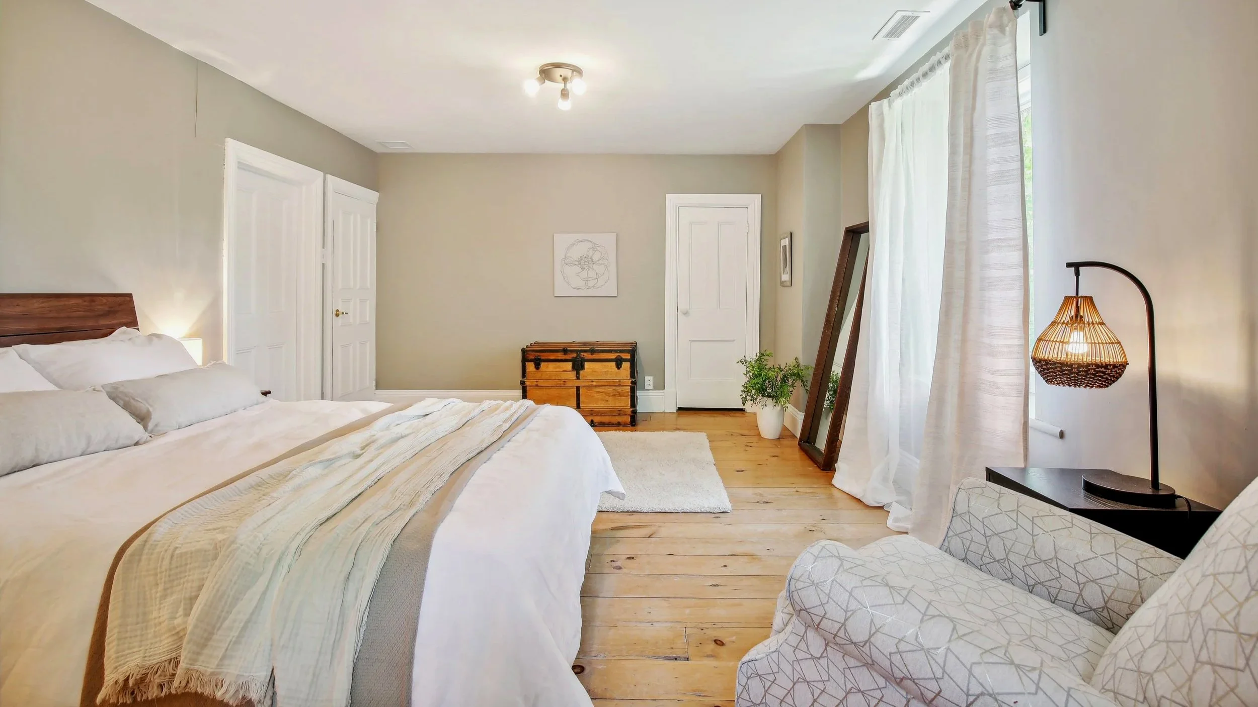 Bright bedroom with hardwood floors, large bed with white bedding and multiple pillows, a plaid armchair, a standing lamp with a woven shade, a mirror leaning against the wall, a chest against the back wall, and large windows with white curtains.
