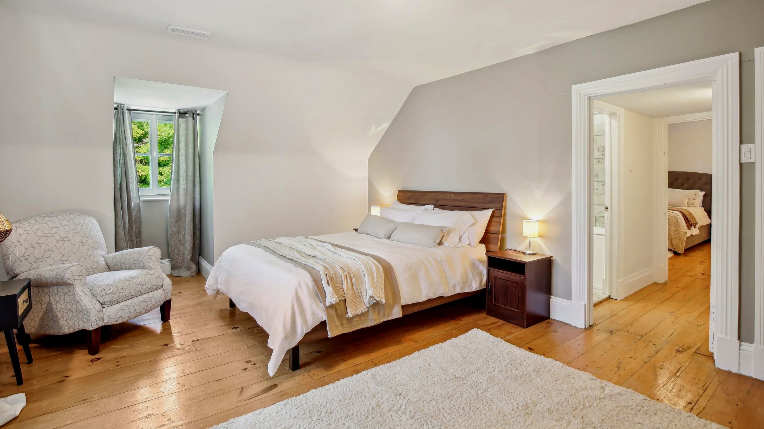 Bedroom with a bed, nightstand with lamp, armchair, window with curtains, and an open door to another bedroom.