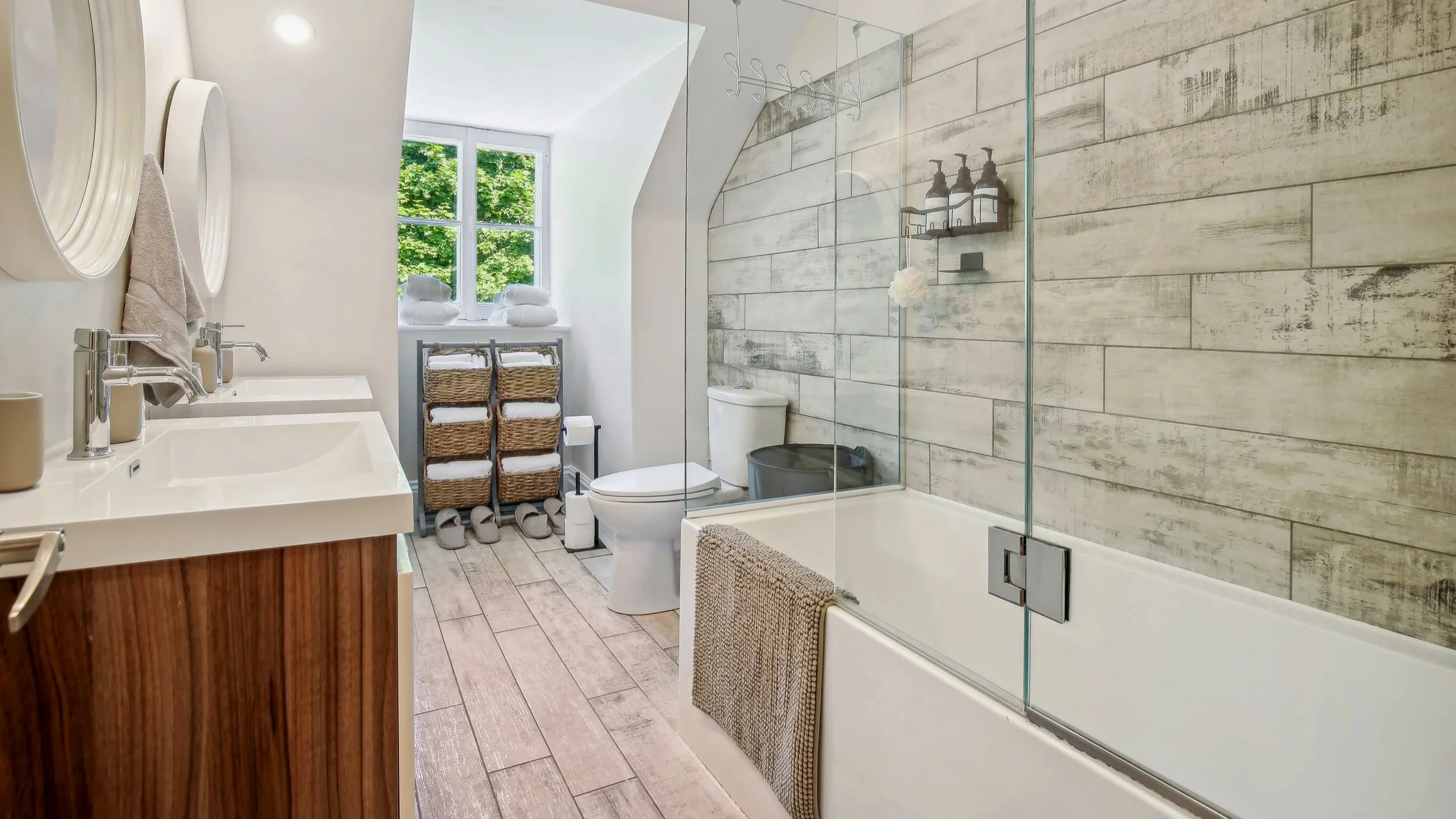 Bathroom with a double sink vanity, wall mirrors, a window with green trees outside, towels, wicker baskets, a toilet, a bathtub with a glass shower enclosure, wall-mounted shelves, and a gray trash bin.