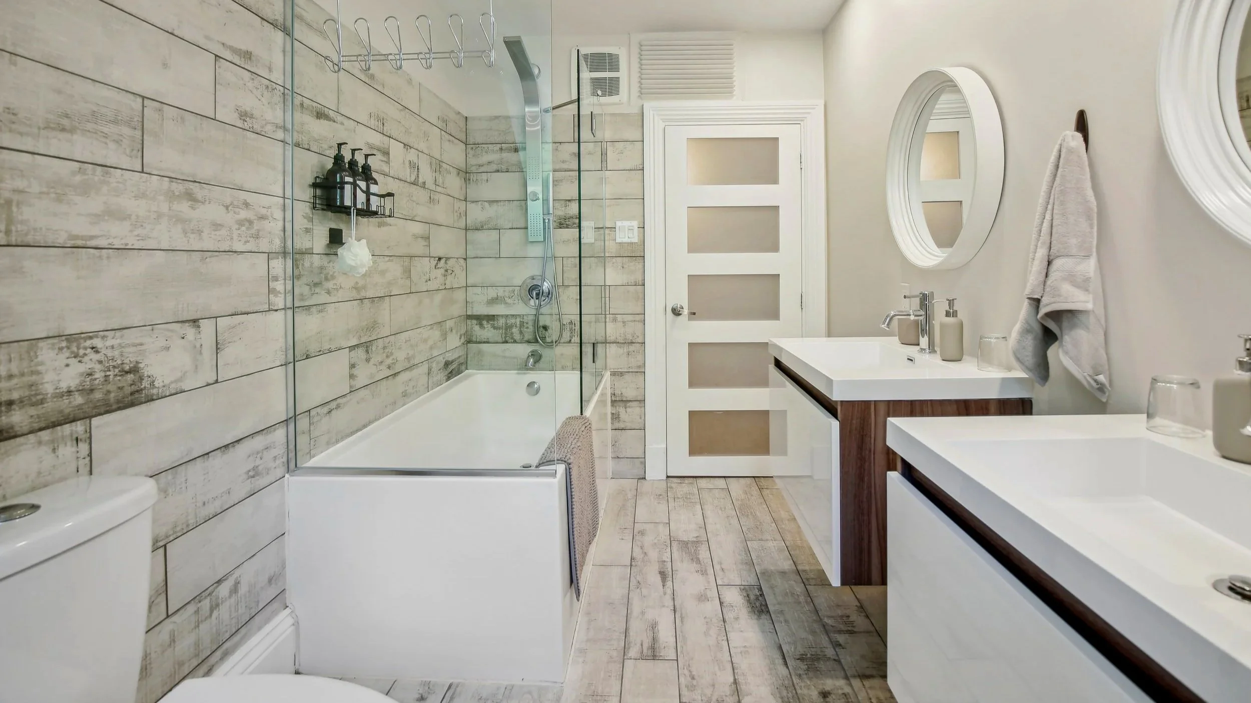 Modern bathroom with double sinks, oval mirrors, a bathtub with a wood-look tile wall, and various toiletries on the counters.