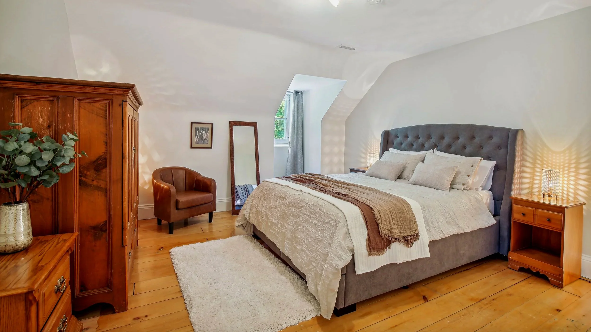 A cozy bedroom with a large bed featuring a gray upholstered headboard, white bedding, and beige pillows. On each side of the bed are matching wooden nightstands with small lamps. To the left, there is a wooden wardrobe, a small wooden dresser with a