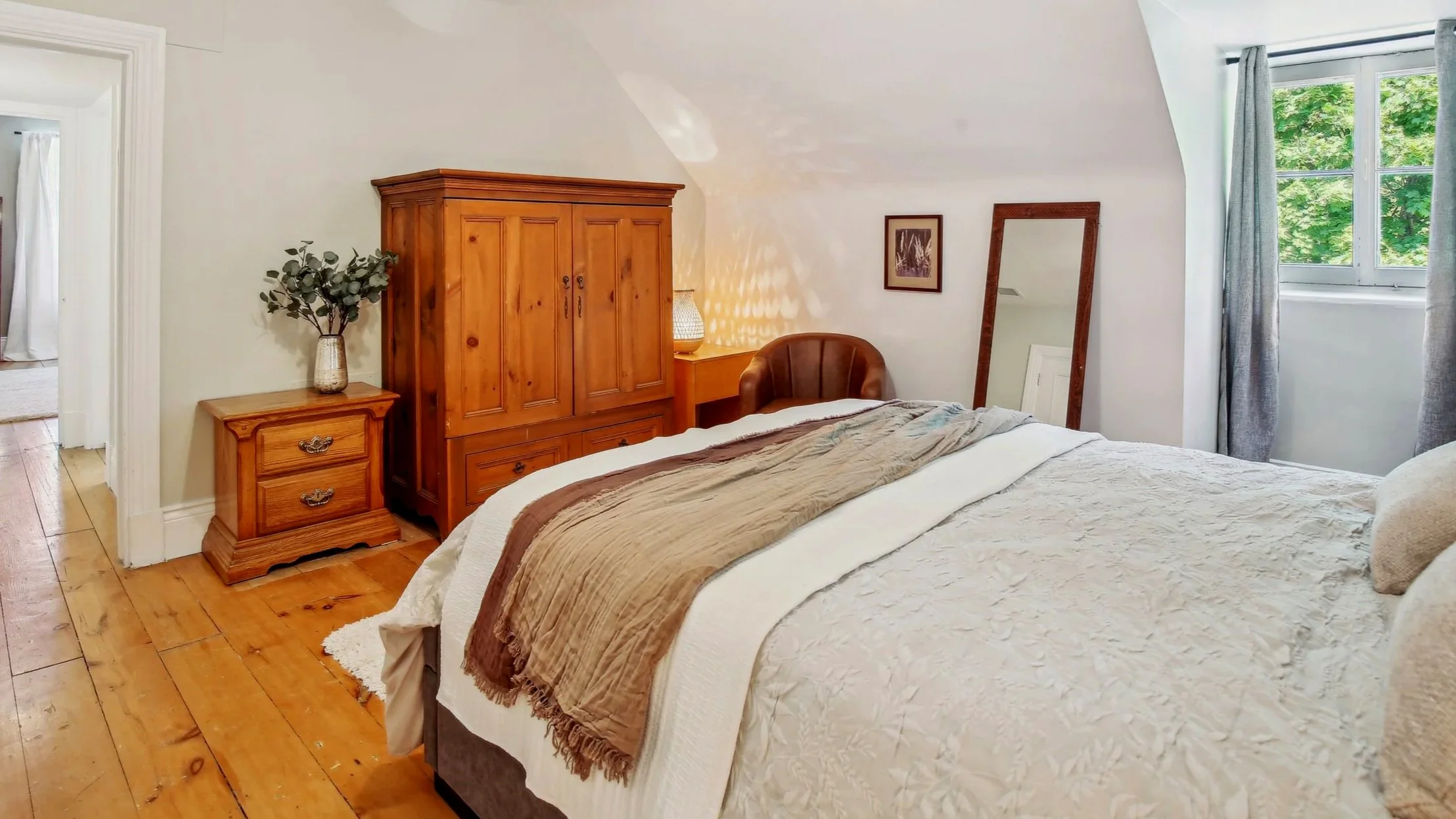 A cozy bedroom with a white textured bedspread, a brown blanket, wooden furniture including a wardrobe, nightstand, and a mirror, as well as a window with gray curtains and green trees outside