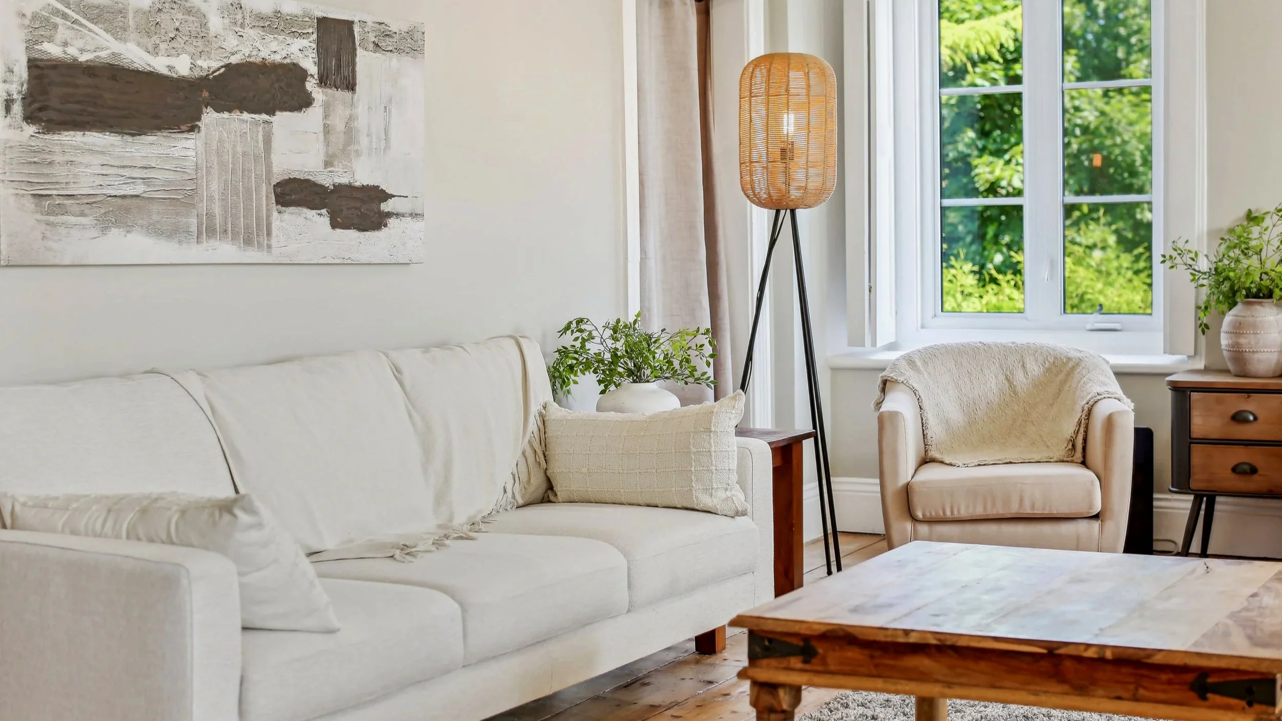 Cozy living room with beige sofa, armchair, wooden coffee table, large window with green trees outside, potted plants, abstract wall art, and a tall floor lamp