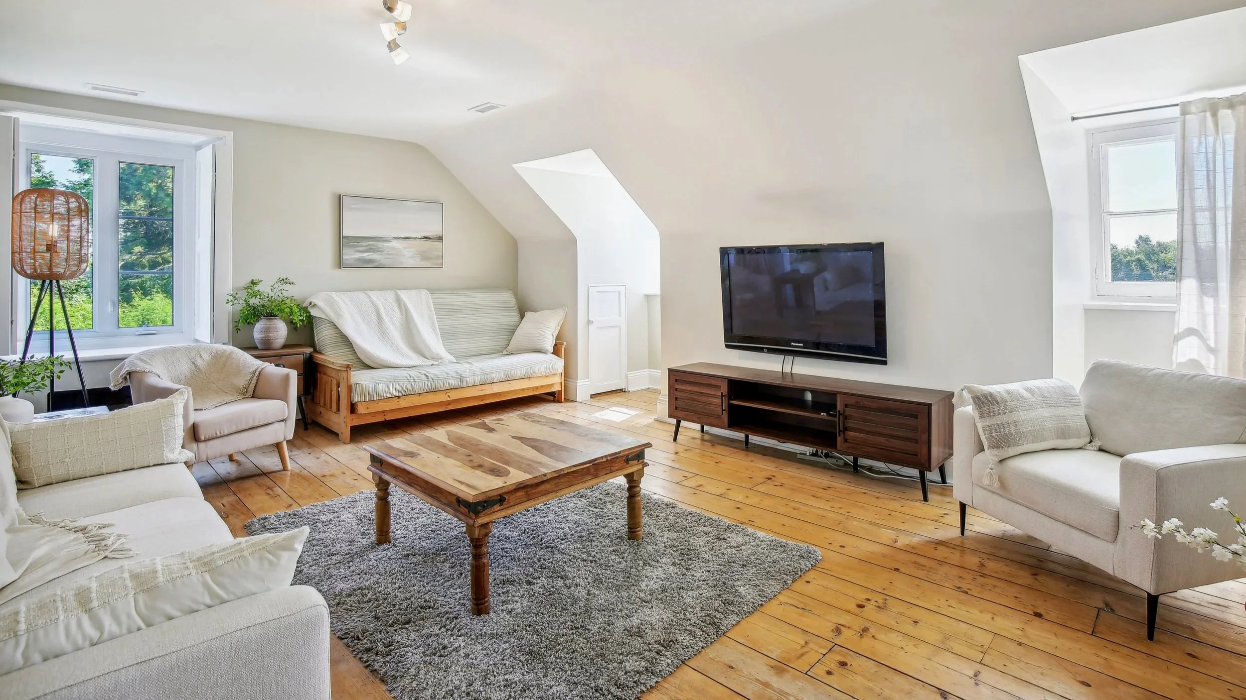 Cozy living room with white walls, wooden flooring, a large flat-screen TV on a wooden stand, white and beige sofas, a wooden coffee table, and large windows letting in natural light, with some greenery visible outside.