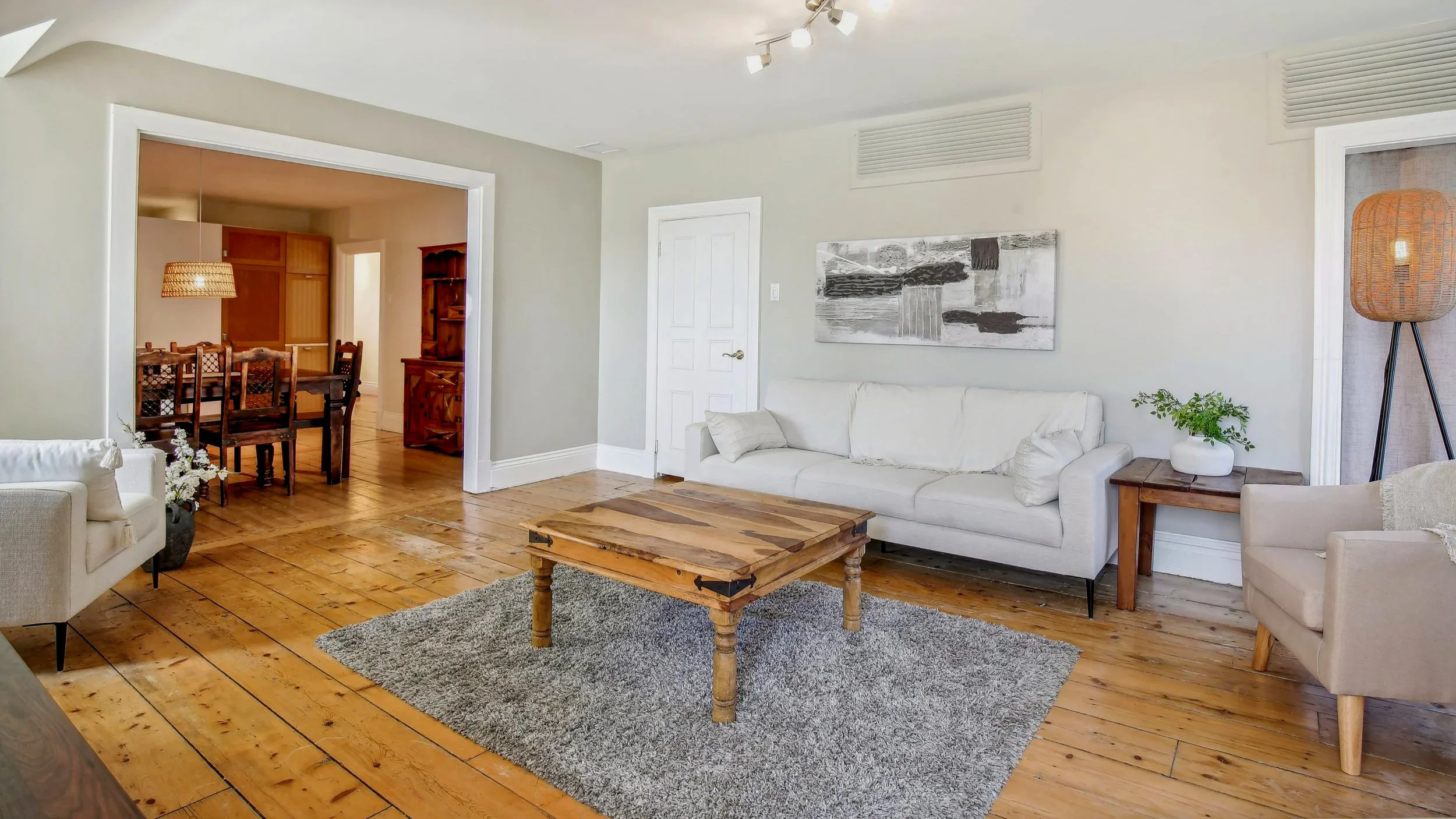 Living room with white sofa, beige armchair, wooden coffee table on gray rug, side table with potted plant, floor lamp, and wall art fixture; adjacent dining area with wooden table and chairs visible through doorway.