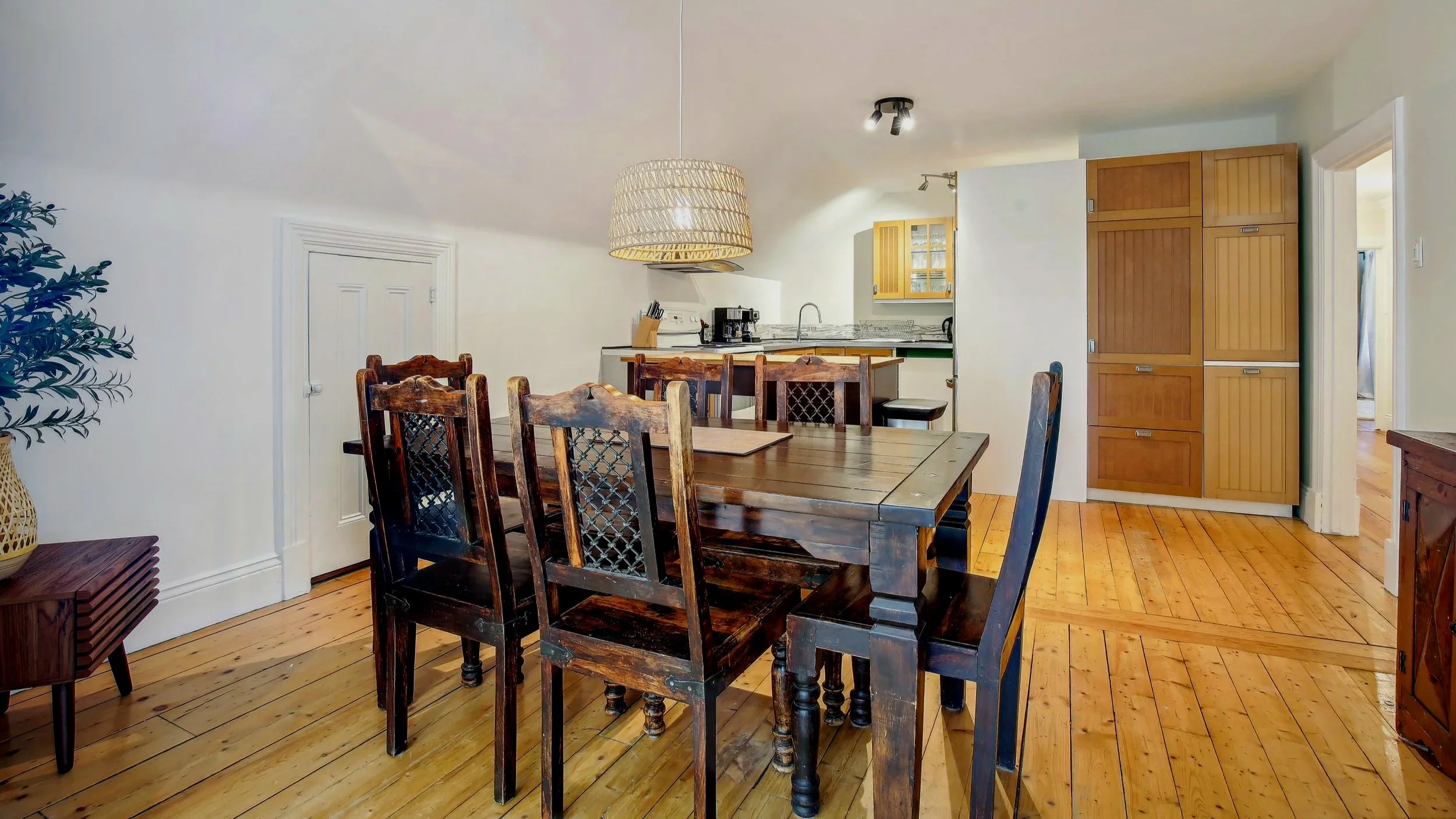 Dining room with wooden table and chairs, open kitchen with wooden cabinets and marble countertops, hardwood floors, and a woven pendant light.