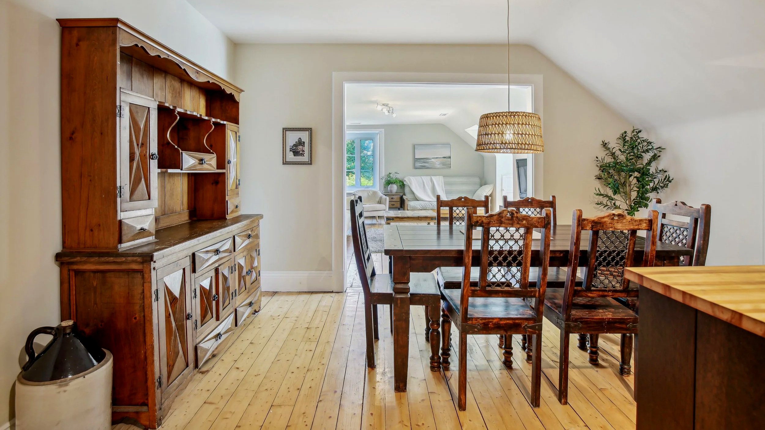 Dining area with a wooden table and six matching chairs, a large wooden buffet against the wall, a hanging wicker lampshade, and a potted plant. In the background, through an opening, a living room with a white couch, side tables, and a window.