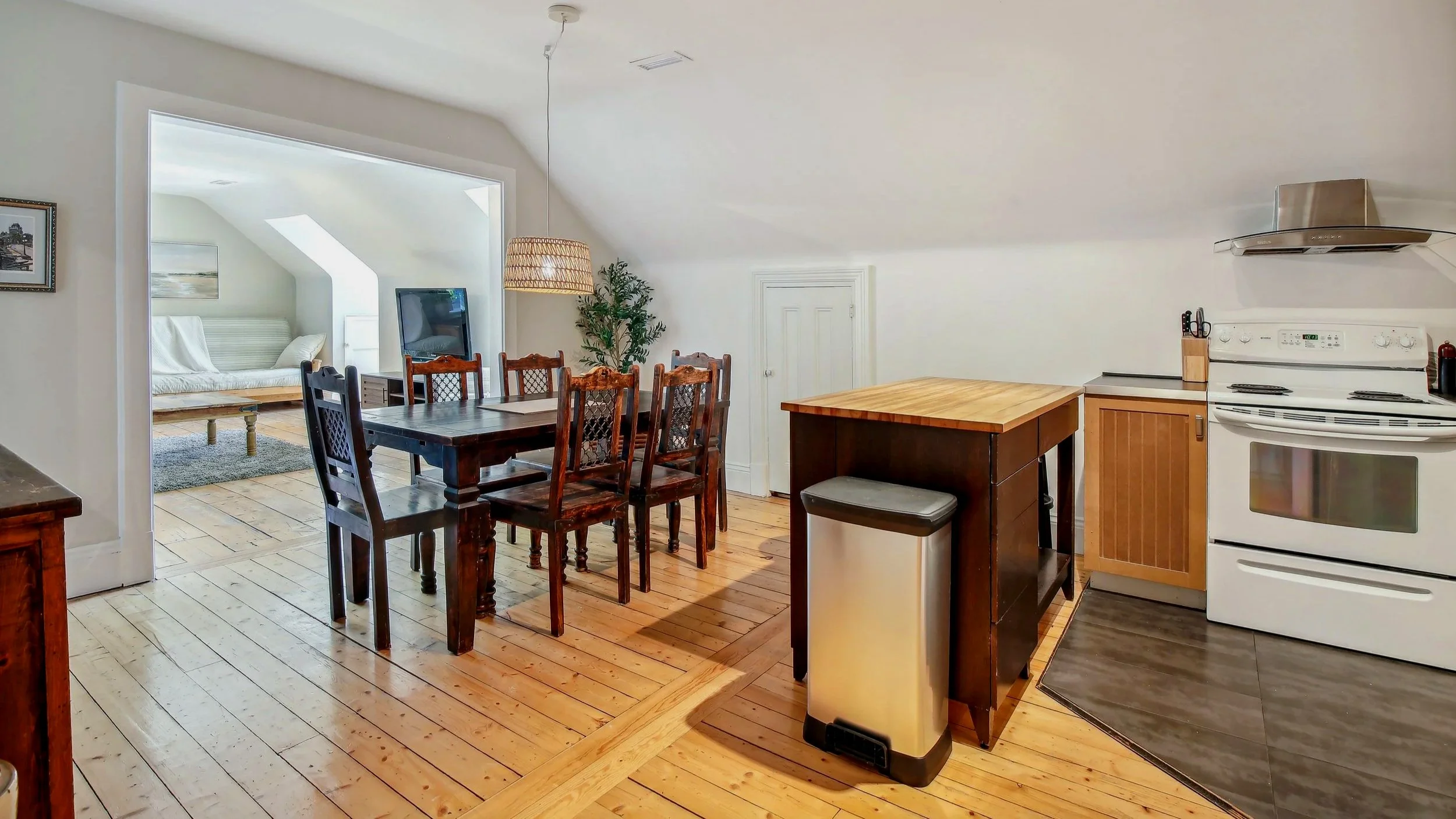 Open-concept kitchen and dining area with wood flooring, white walls, a wooden dining table with six chairs, a kitchen counter, stove, and a small living space with a sofa and TV in the background.