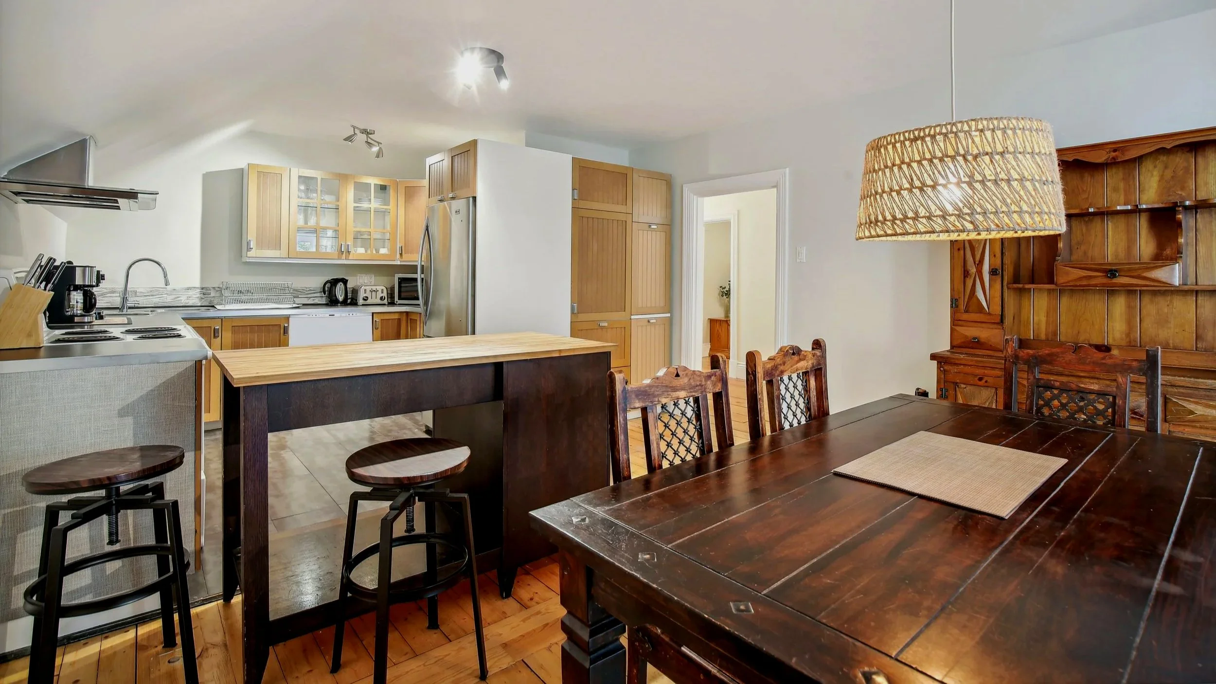 Kitchen and dining area with wooden furniture, a dark wood dining table, and kitchen cabinets; kitchen has a white refrigerator and counter appliances; large wooden hutch and a wicker lampshade hanging over the table.