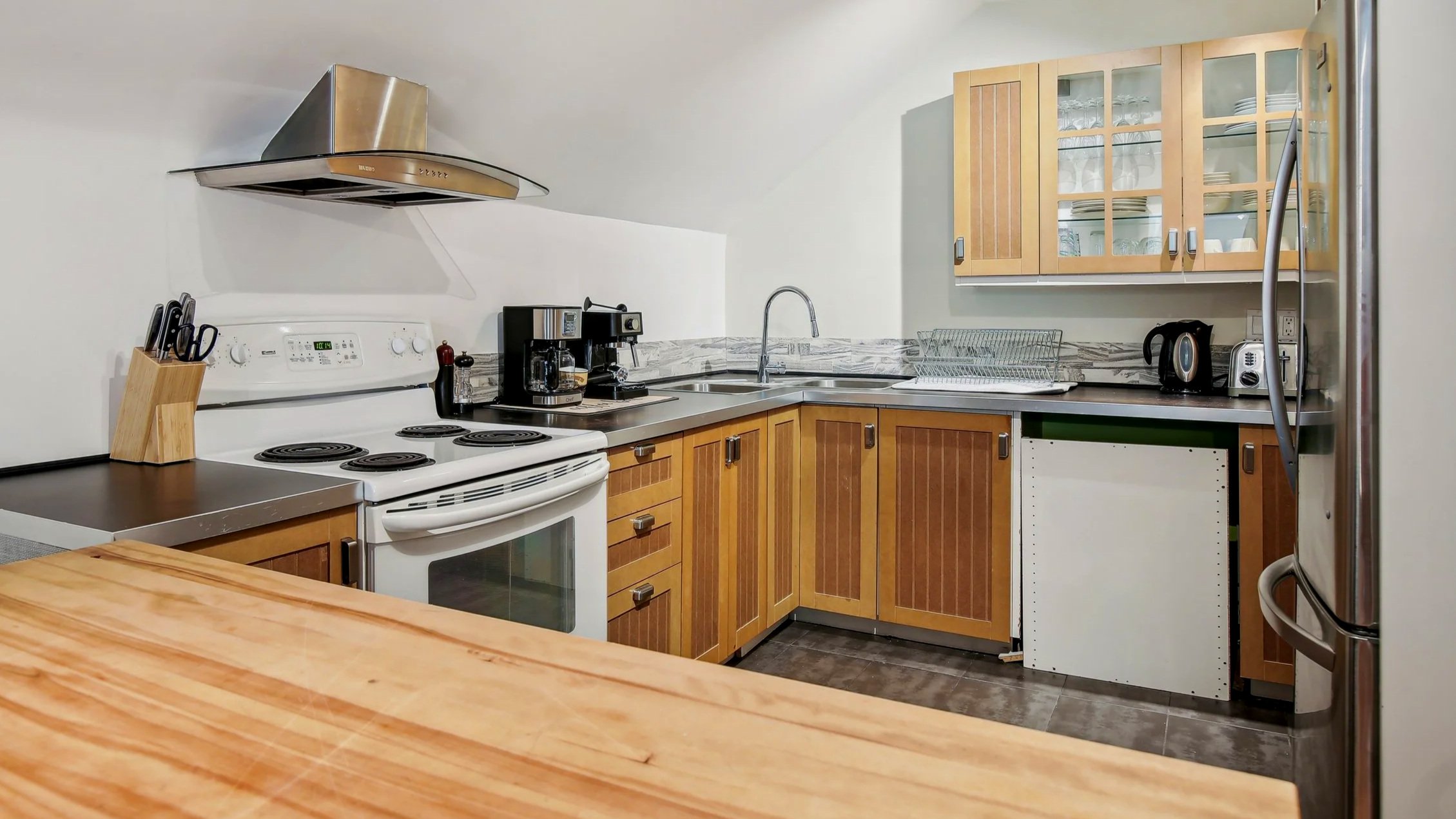 Kitchen with white stove, wooden cabinets, stainless steel range hood, coffee maker, dish rack, refrigerator, and various small appliances.