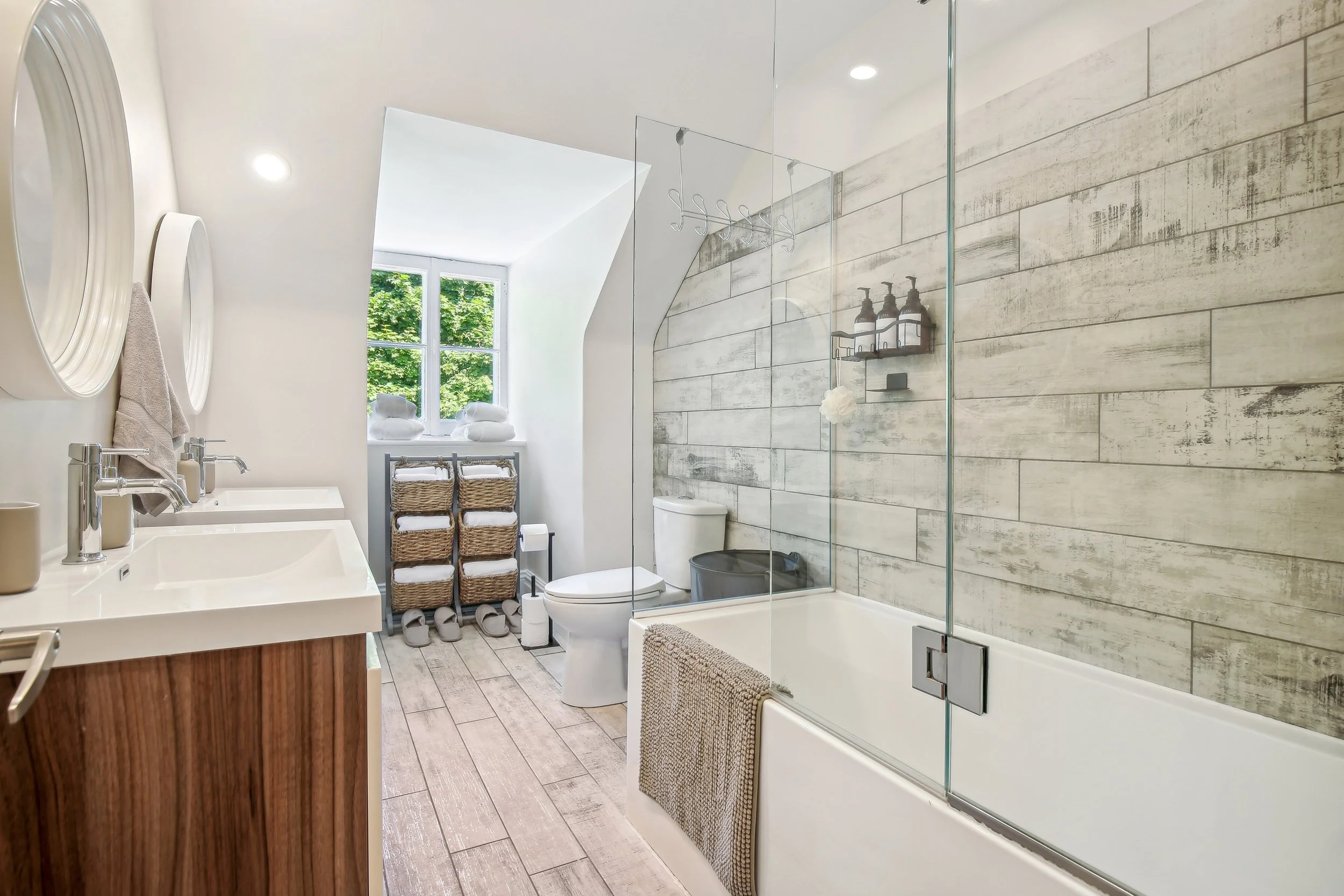 A modern bathroom with a wooden vanity, white sink, large window, and a walk-in shower with glass panels. There are baskets and towels near the window, and the walls are decorated with light-colored wood-like tiles.