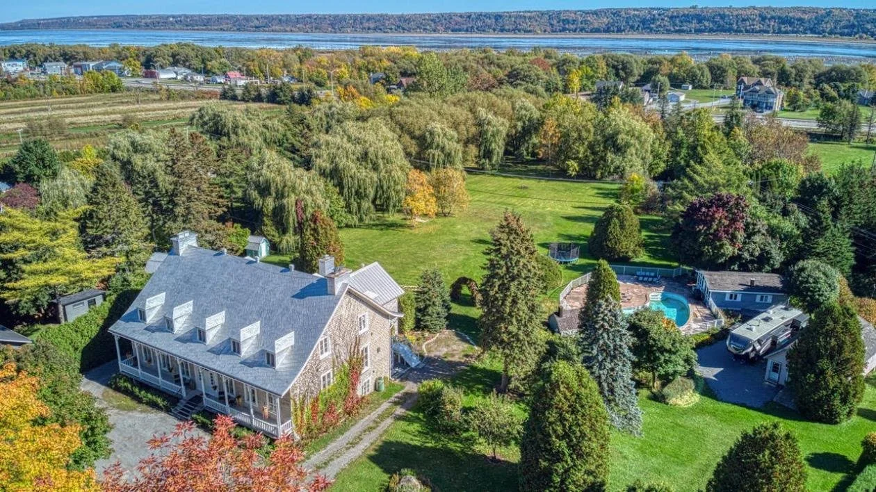 Aerial view of a large house with a gray roof surrounded by lush green trees, a swimming pool with a fence, and a well-maintained lawn. In the background, there is a body of water and a small town.