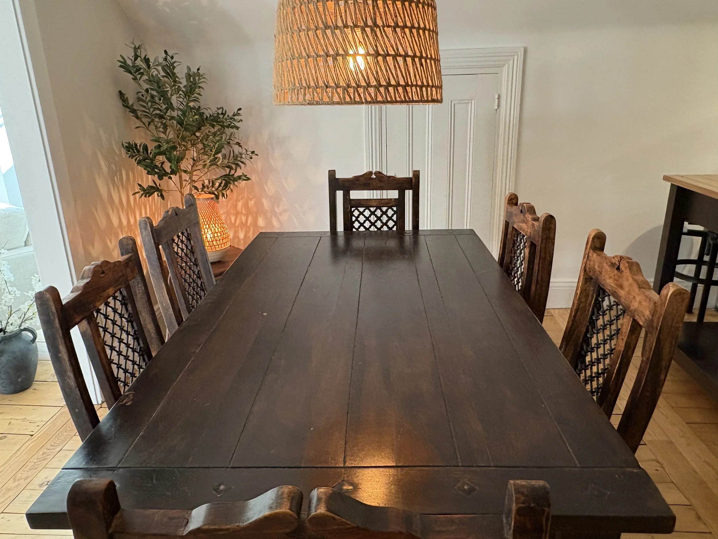A wooden dining table with six matching wooden chairs, a wicker pendant lamp overhead, a potted plant on the left, and a sideboard on the right in a room with hardwood floors and light-colored walls.