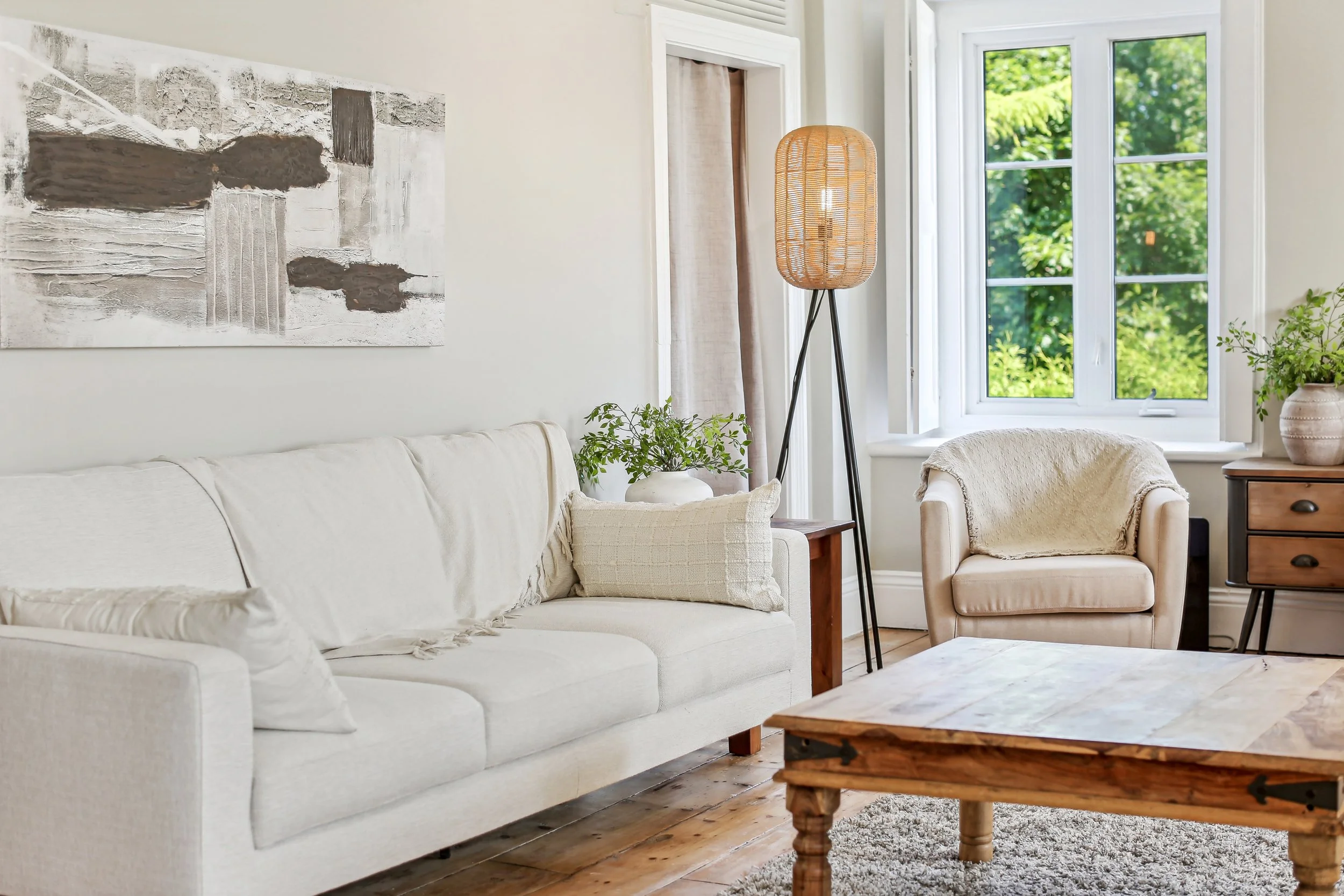 Living room with a white sofa, a matching armchair, wooden coffee table, artwork on the wall, potted plants, a standing floor lamp, and a window with greenery outside.