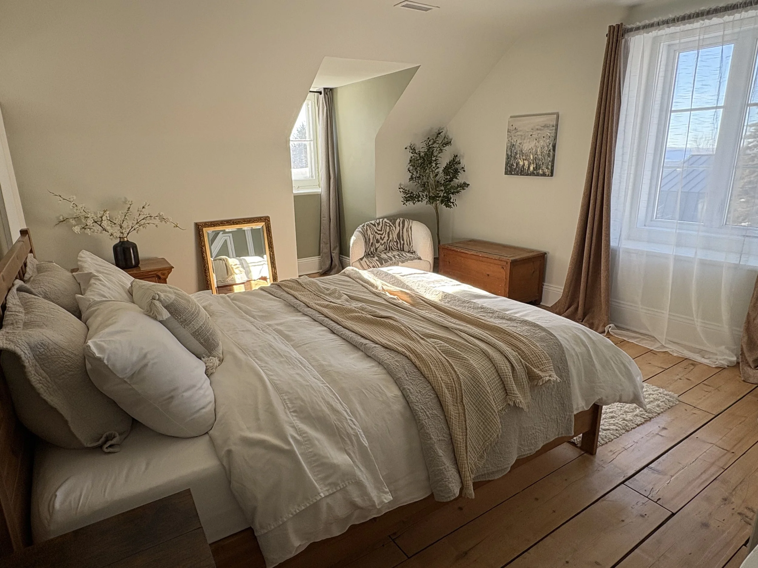 A cozy bedroom with a large bed featuring white bedding, decorative pillows, and a beige throw blanket. There is a side table with a vase of white flowers, a mirror, a chair with a zebra print cushion, a wooden chest, and a window with sheer white cu