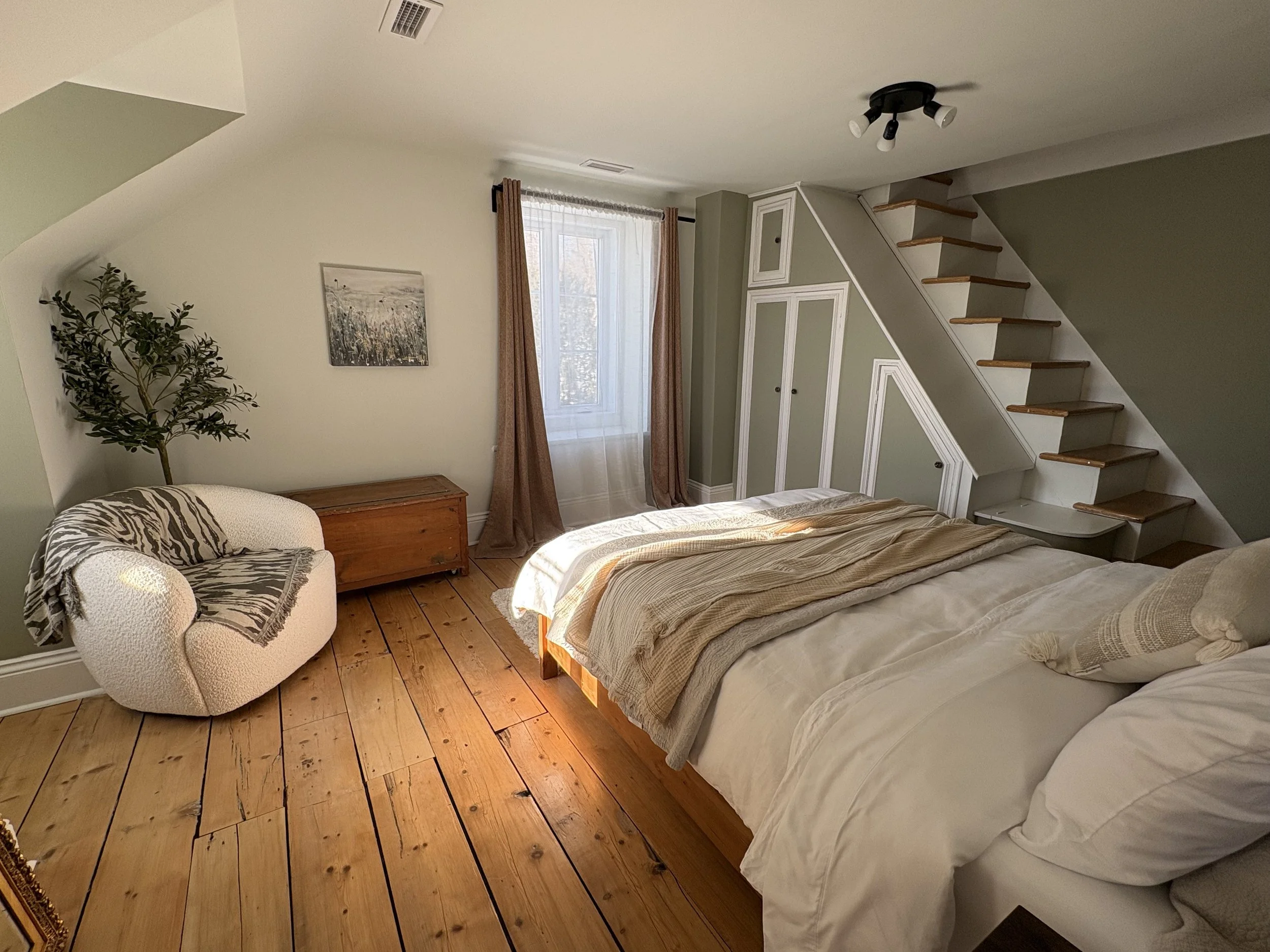 A bedroom with a wooden floor, a bed with light-colored bedding, a small armchair with a zebra-patterned blanket, a window with curtains, a built-in closet, and a staircase leading upwards.