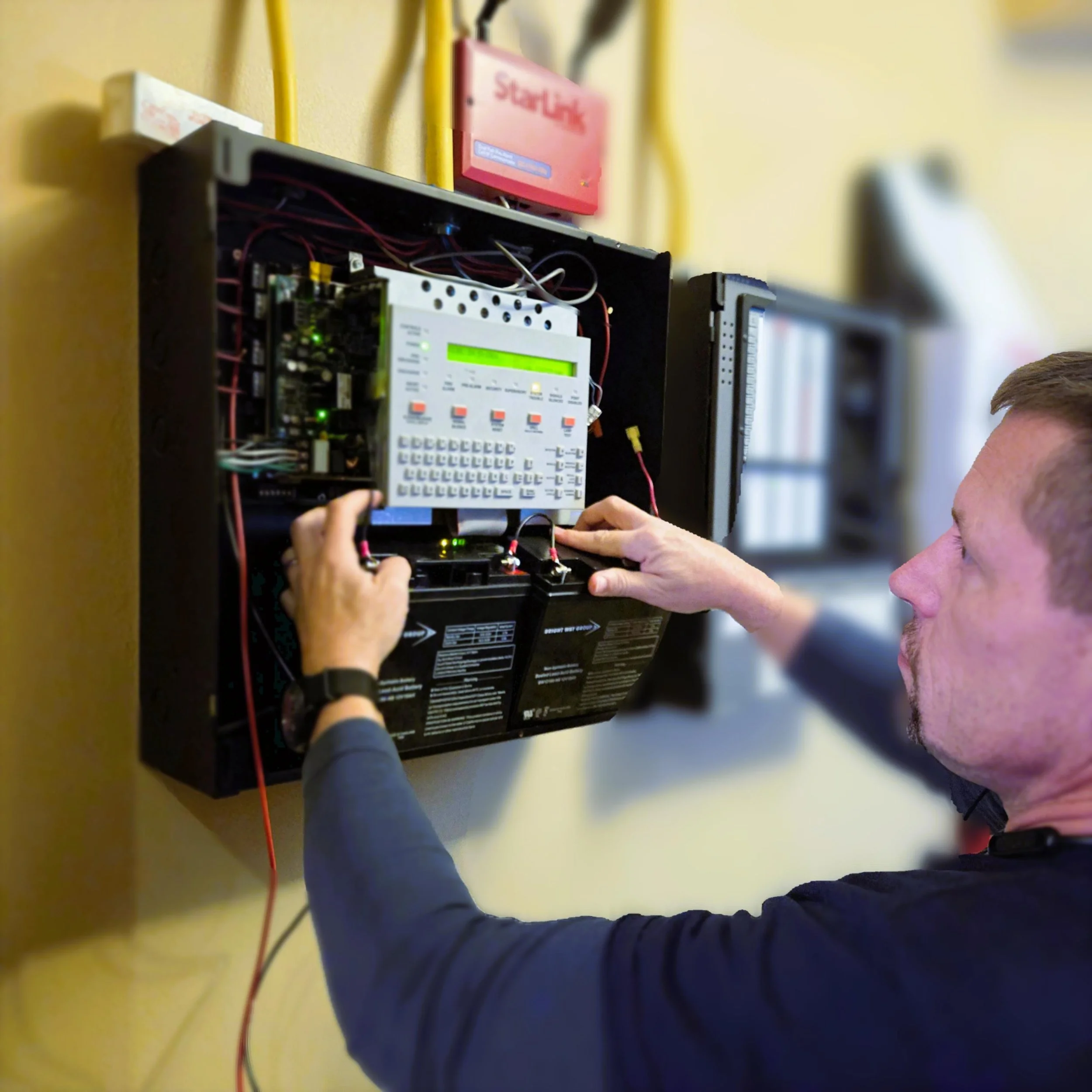 Man working on an open electrical panel with wires and electronic devices inside, mounted on a yellow wall.