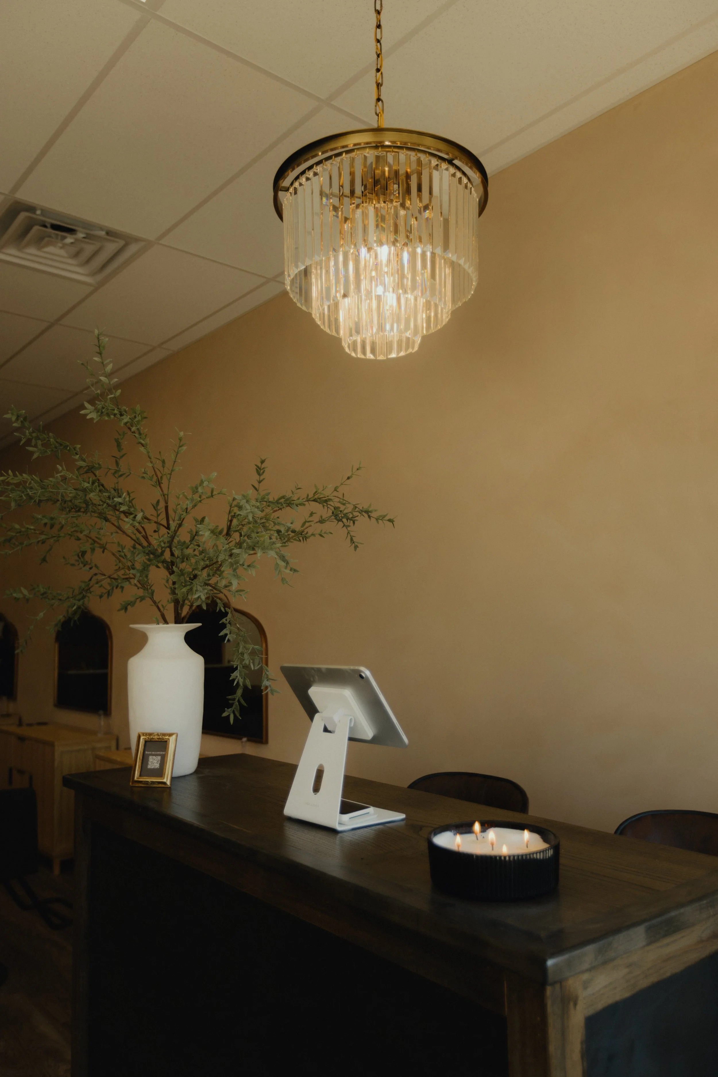 Indoor scene featuring a decorative hanging chandelier with crystal elements illuminating a wooden table. The table has a large white vase with greenery, a small picture frame, a white electronic device, and a black candle holder with lit candles. The background has a beige wall with a plant in the corner.