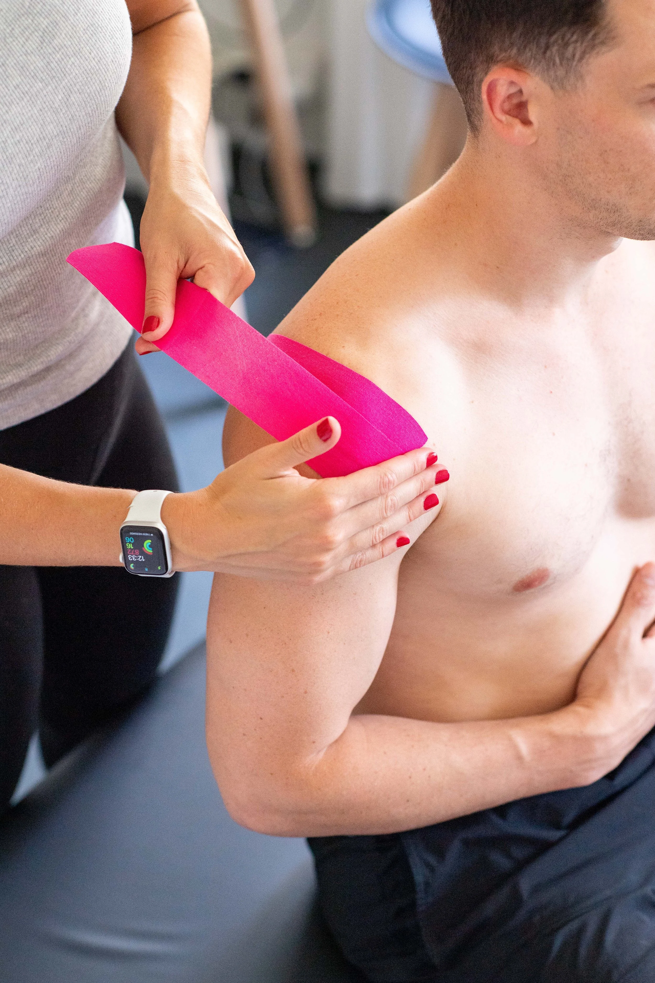 A therapist or trainer is taping a man's shoulder with pink kinesiology tape.