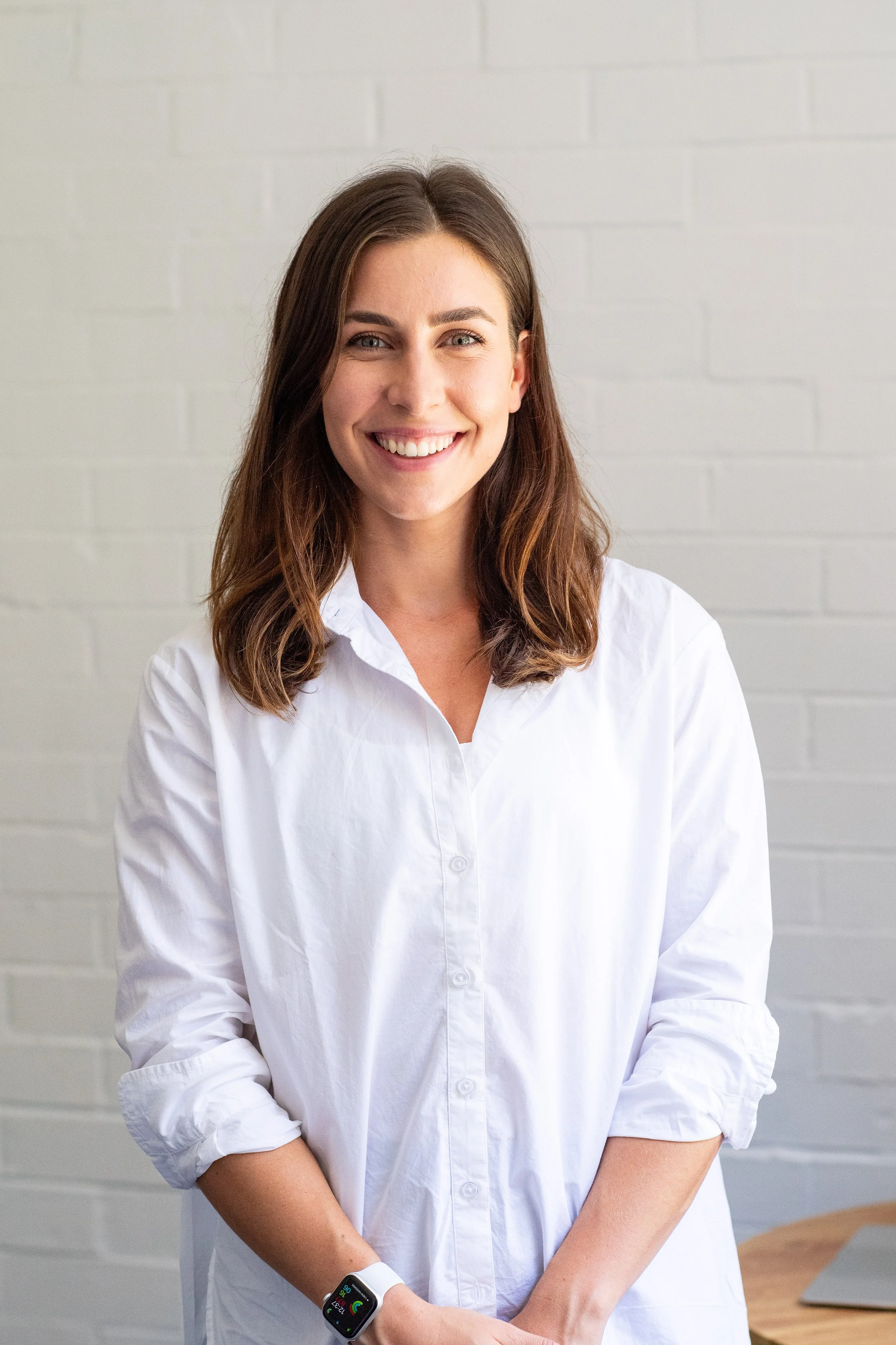 A woman with shoulder-length brown hair, smiling, wearing a white button-up shirt, standing in front of a light-colored brick wall.