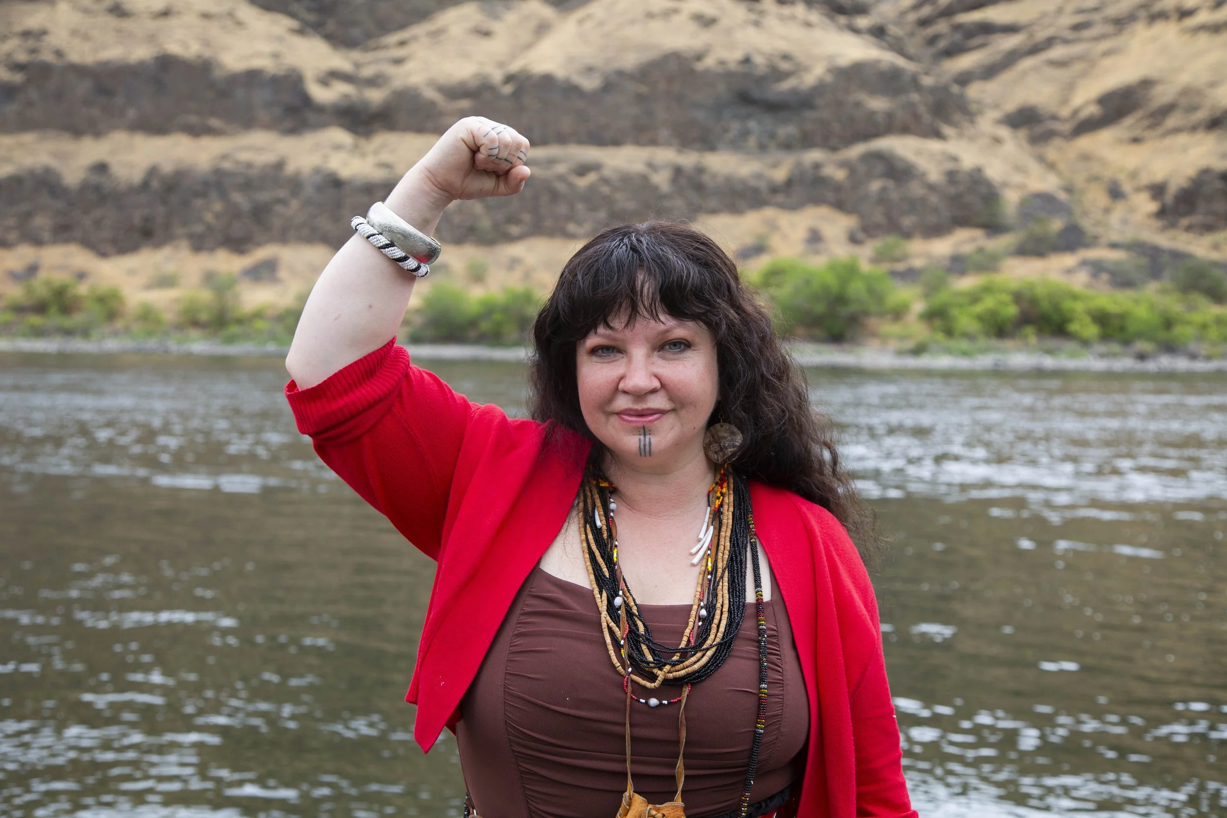 Woman with dark curly hair and face paint, wearing a red jacket, brown top, and layered necklaces, flexing her right arm near a river with mountains and trees in the background.