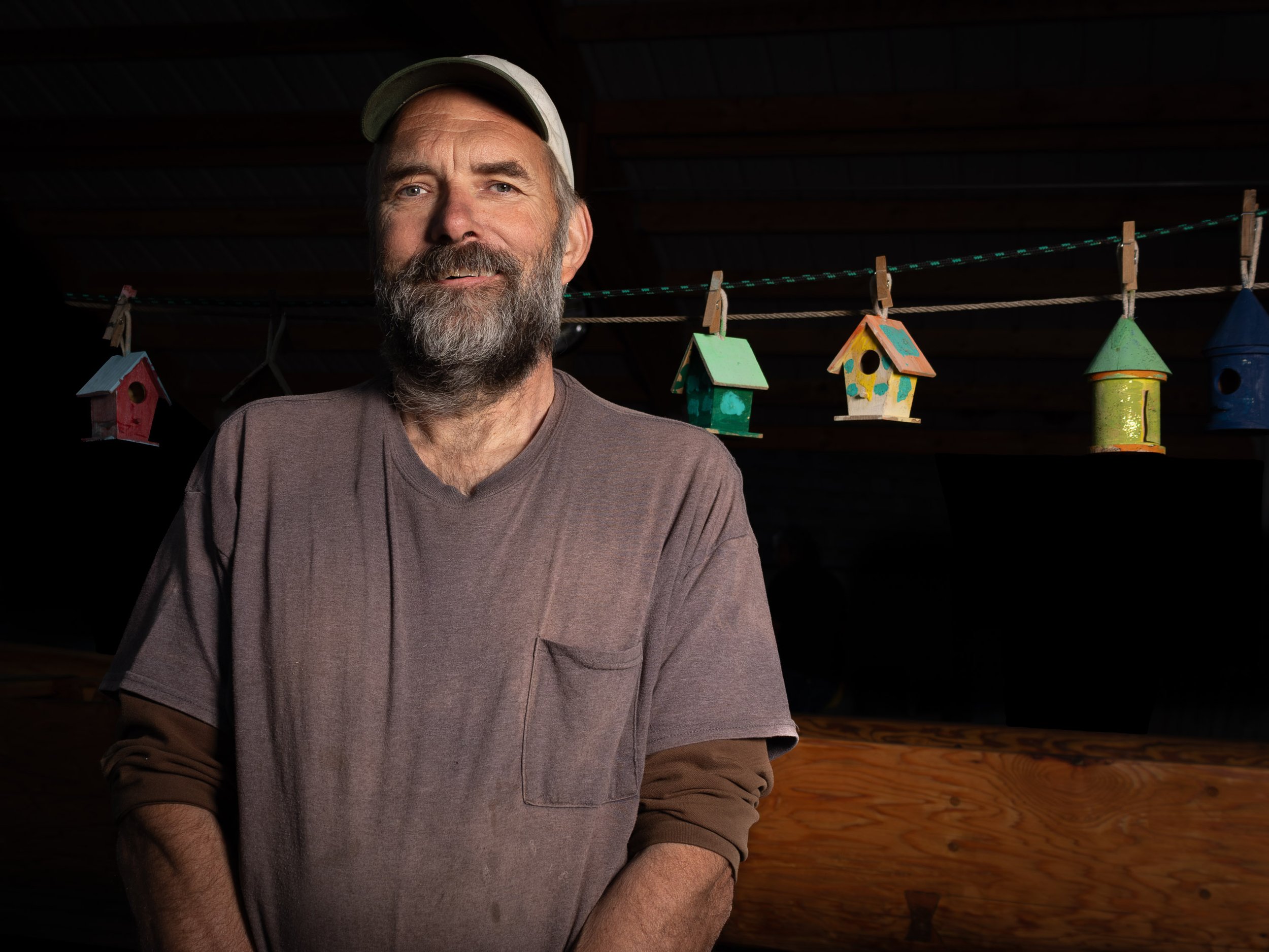 A middle-aged man with a gray beard and mustache wearing a gray baseball cap and a brownish-gray shirt, standing indoors with a dark background and colorful birdhouses hanging on a line behind him.