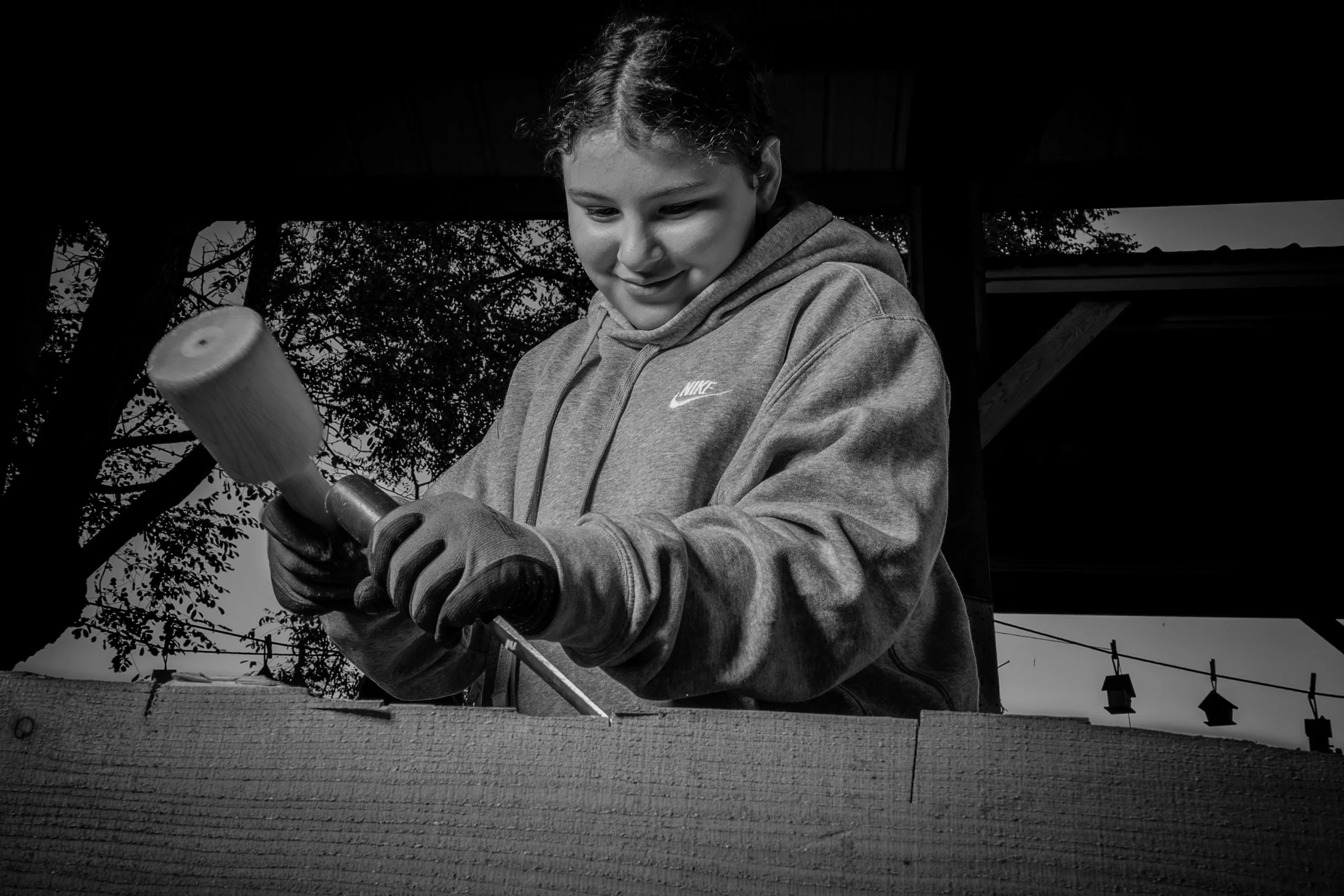 A young person in a hoodie and gloves using a hammer to drive a nail into a wooden surface outdoors, with trees and a shed visible in the background.