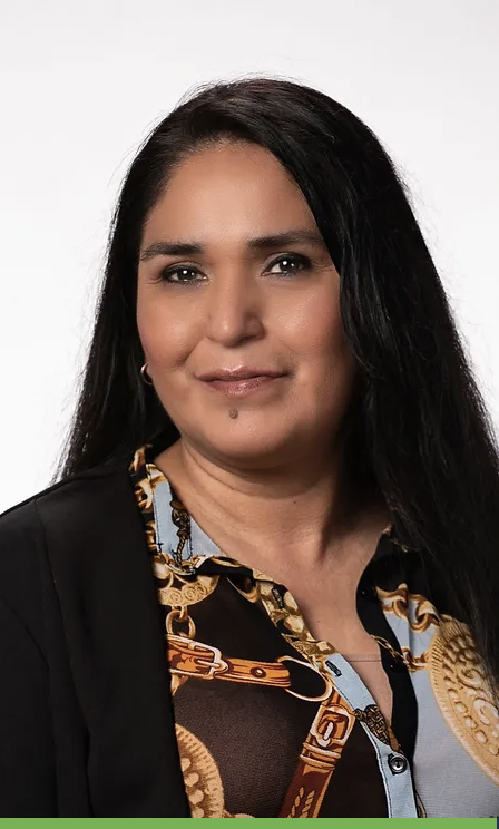A woman with long dark hair, wearing a black blazer over a patterned shirt, smiling slightly against a plain white background.