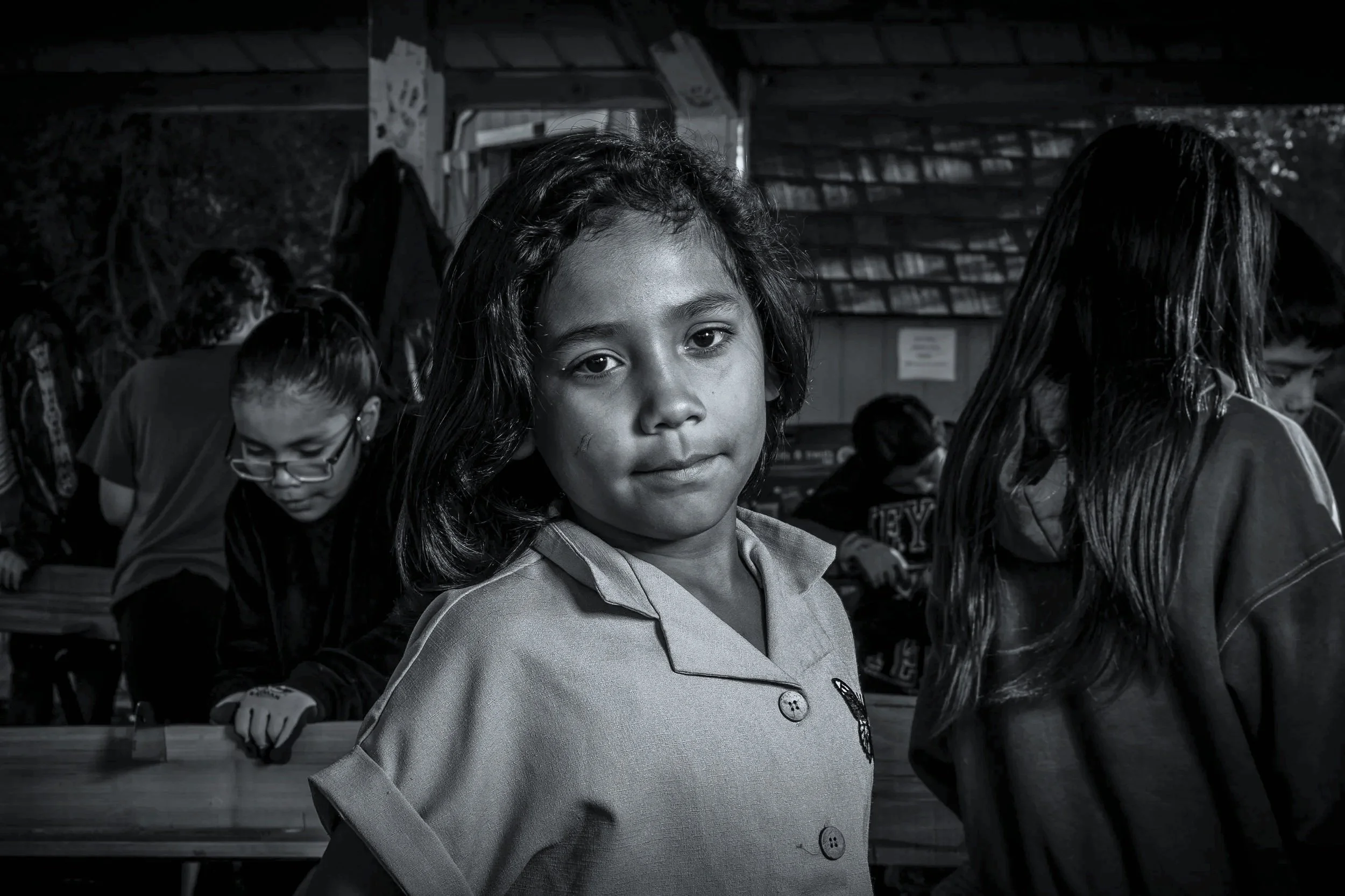 A young girl with dark hair and a serious expression standing in a classroom, surrounded by children engaged in activities, with bookshelves in the background.