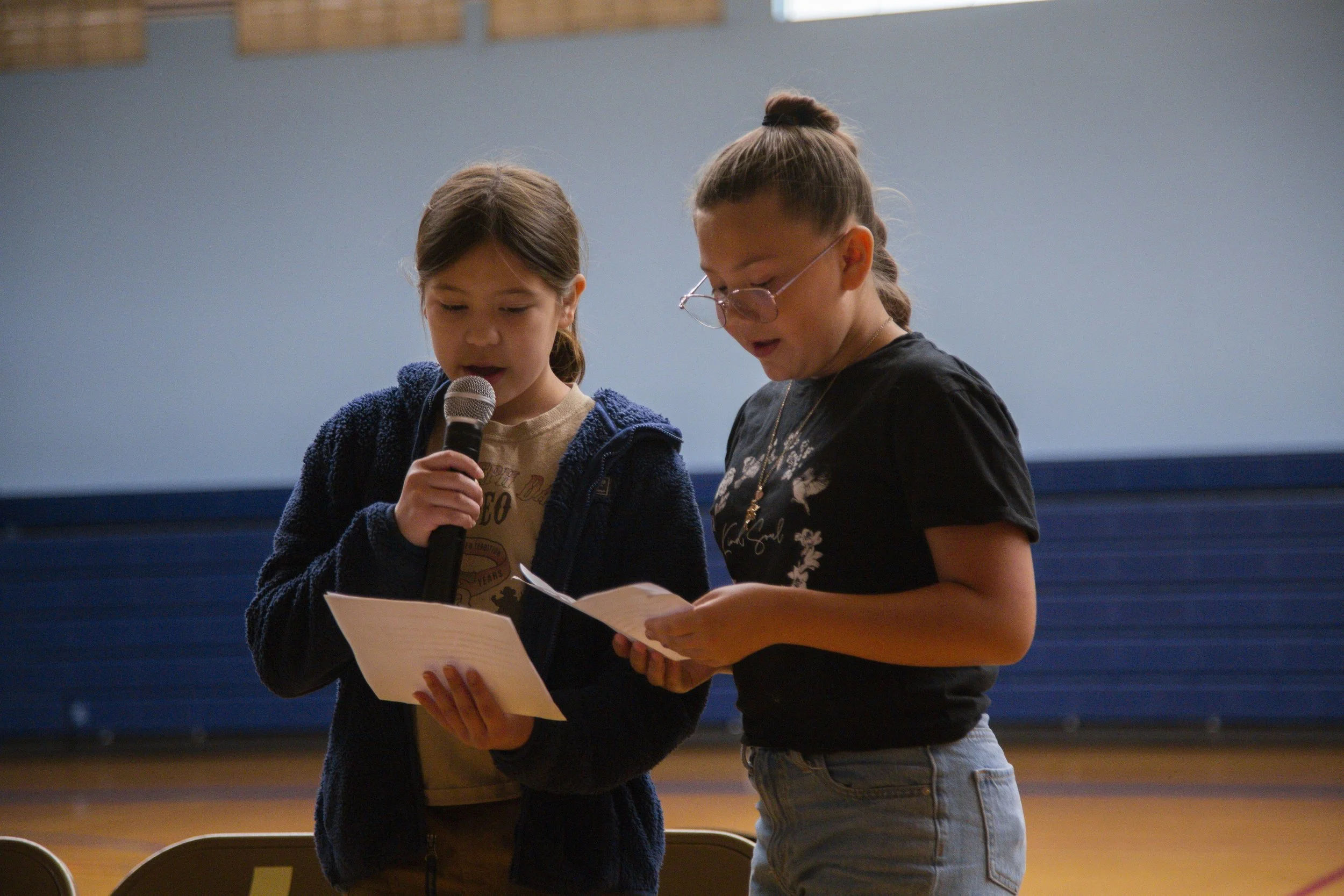 Two young girls standing close together, reading from papers and speaking into a microphone in an indoor setting, possibly a school gym or auditorium.