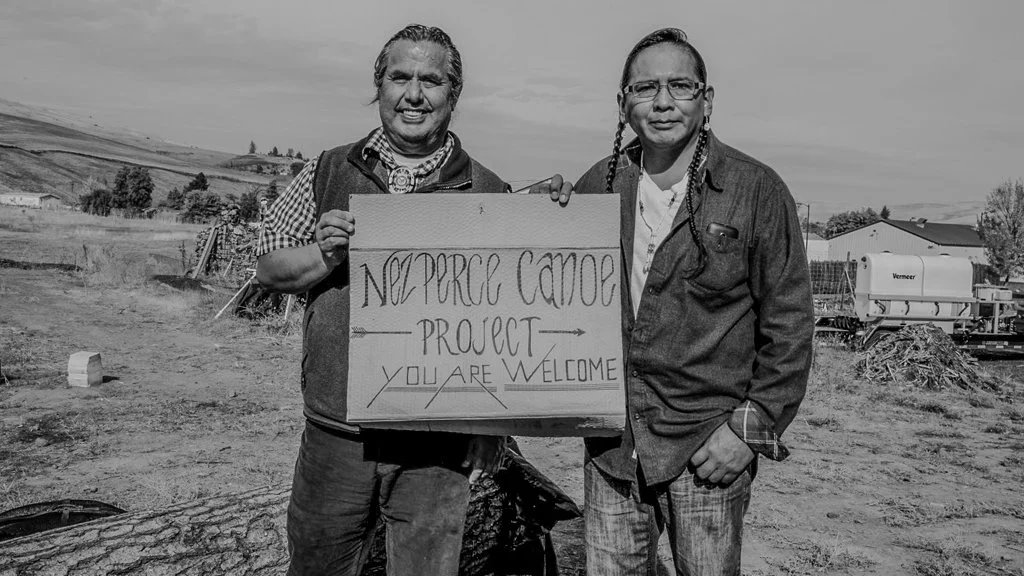 Two indigenous men standing outdoors, holding a sign that reads "NO PER CAPE CAME PROJECT YOU ARE WELCOME" in a rural area with hills and farm equipment in the background.