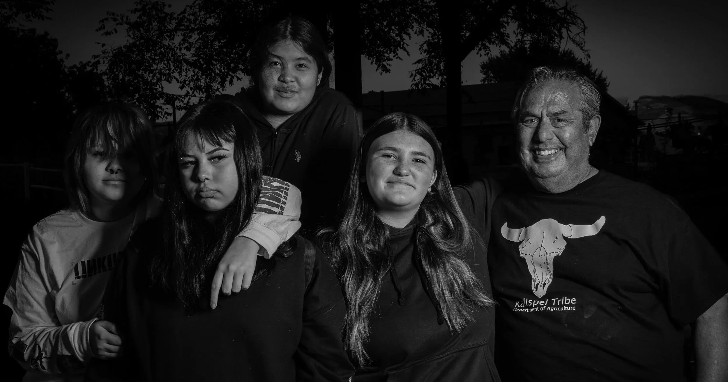 Group of five people standing outdoors at dusk, smiling and posing for the camera. Four are women and one is a man, with trees and a fence in the background. The man is wearing a t-shirt with a longhorn skull and the words "Kalispel Tribe Department of Agriculture." The women have diverse hair styles and expressions.