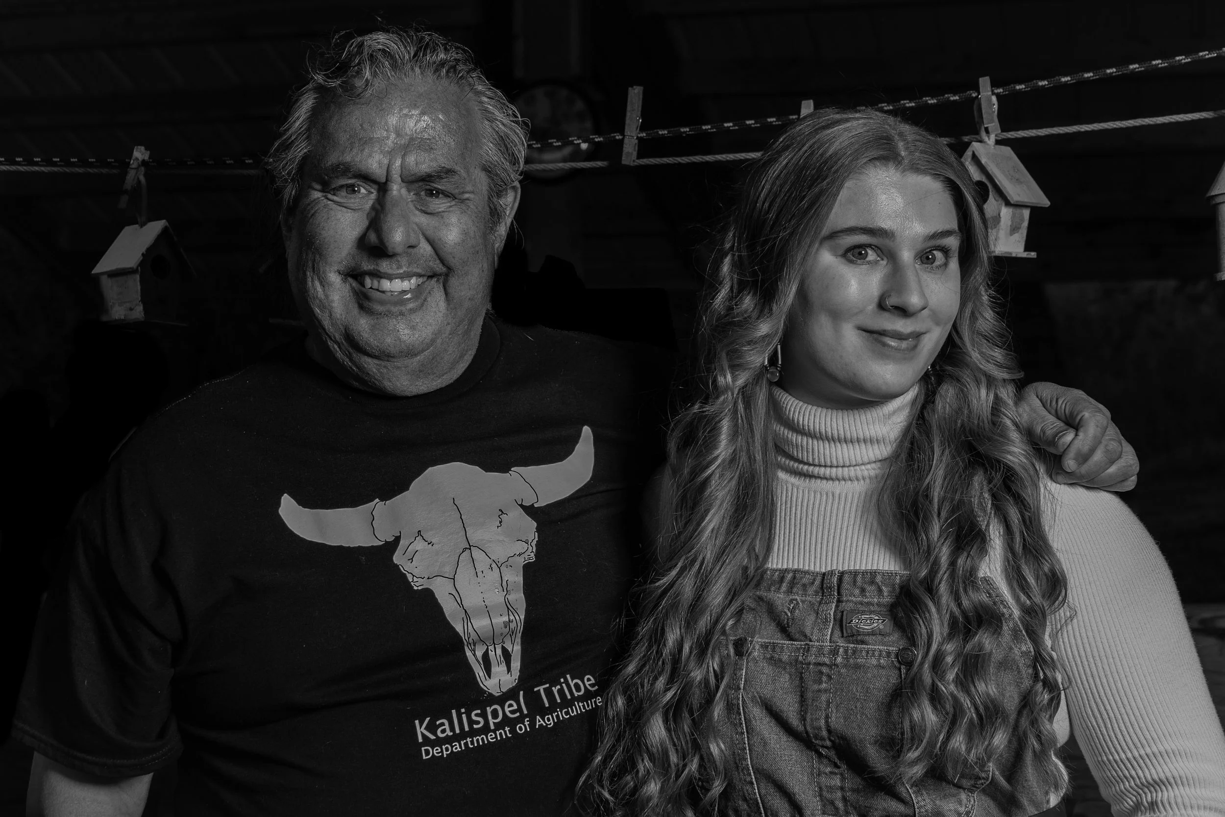 A black and white photo of an older man and a young woman side by side, smiling. The man has long gray hair and is wearing a black t-shirt with a graphic of Texas and the words "Kalispe Tribal" and "Department of Agriculture". The young woman has lon