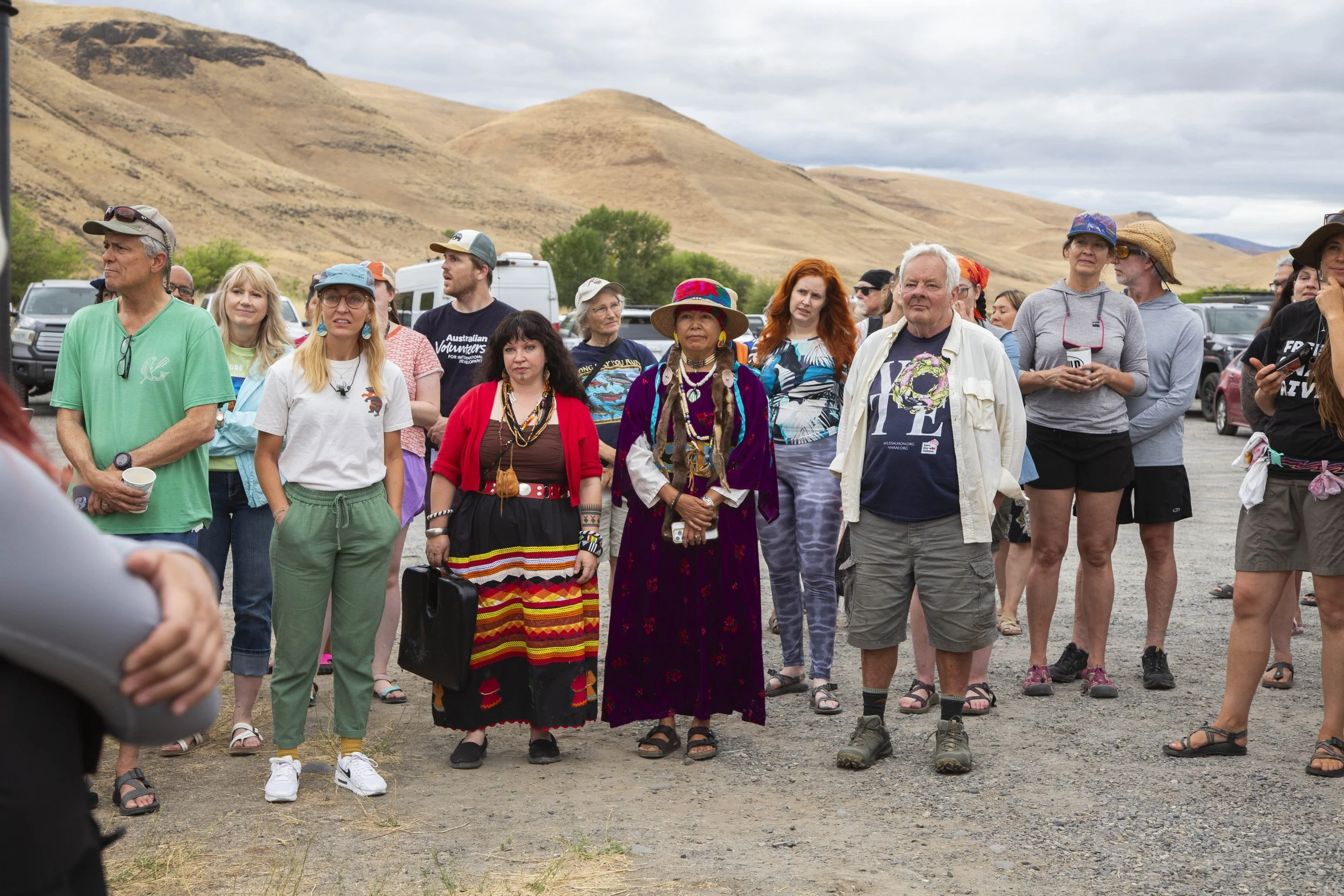 A diverse group of people standing outdoors in a mountainous landscape, some wearing colorful and traditional clothing, others in casual attire, appearing to listen or watch something out of view.