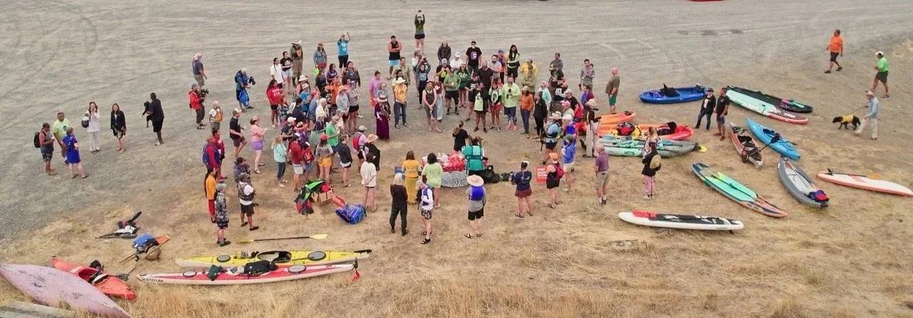 Large group of people gathered on a sandy beach, surrounding multiple colorful kayaks and paddleboards between them.
