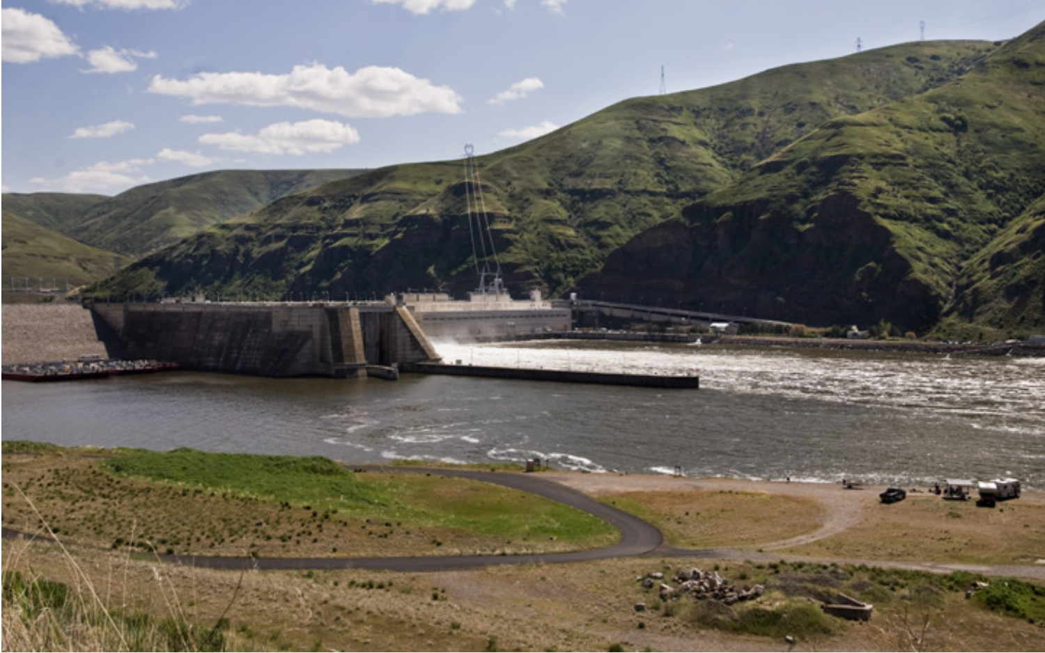 A large dam with water flowing over the edge, situated in a valley surrounded by green hills under a partly cloudy sky.
